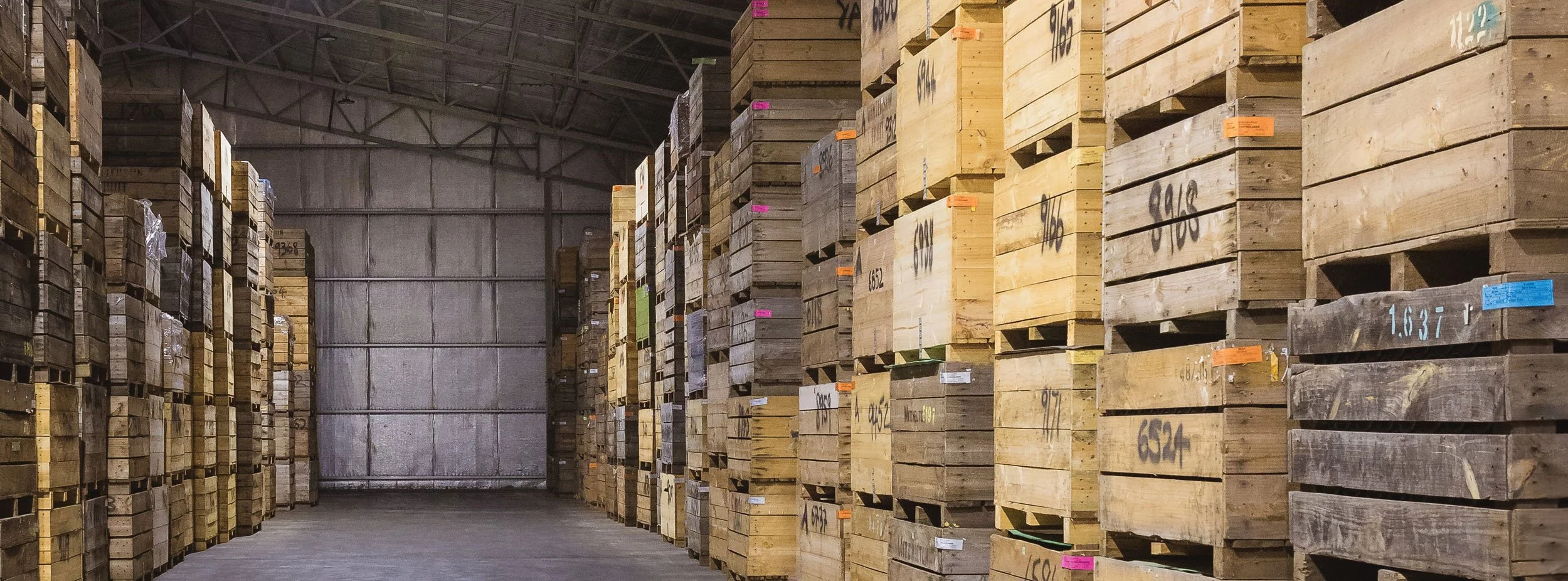 Kilchurn sparkling winemakers warehouse aisle lined with tall stacks of wooden crates on pallets, with some colorful labels and numbers written on the crates.
