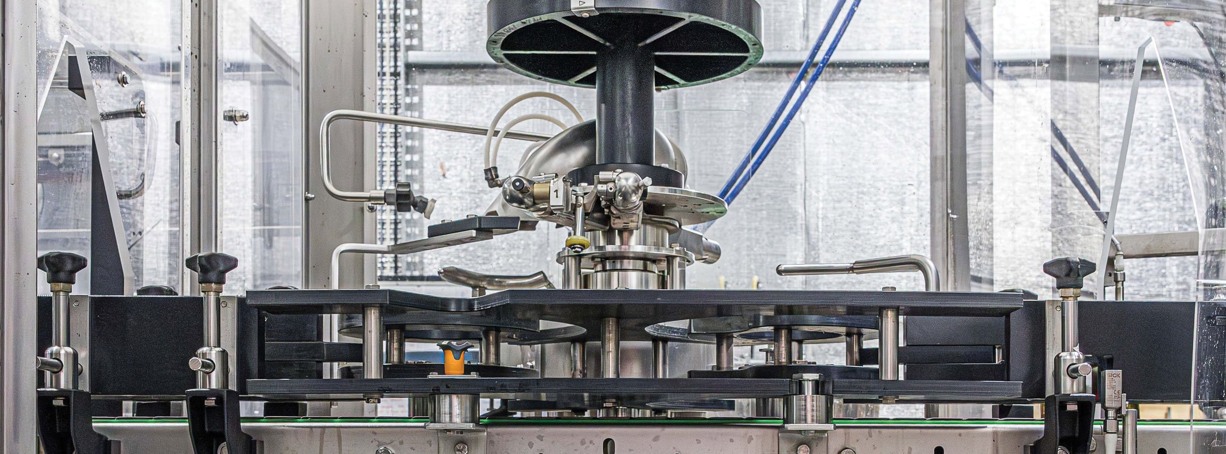 Close-up of a stainless steel laboratory or industrial machine with various pipes, valves, and control knobs, enclosed in transparent safety glass.