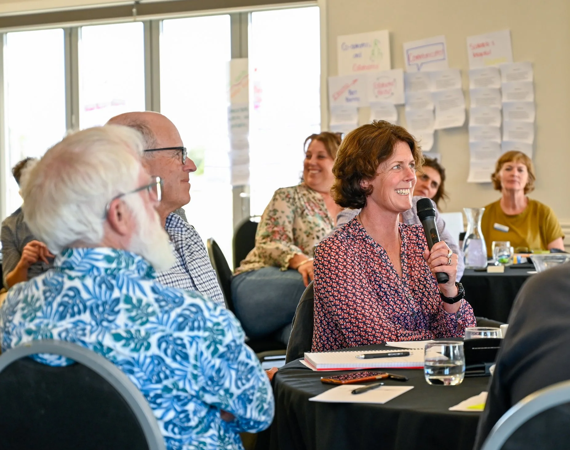 A woman in a patterned blouse speaking into a microphone during a meeting, with several people seated around a table and smiling.