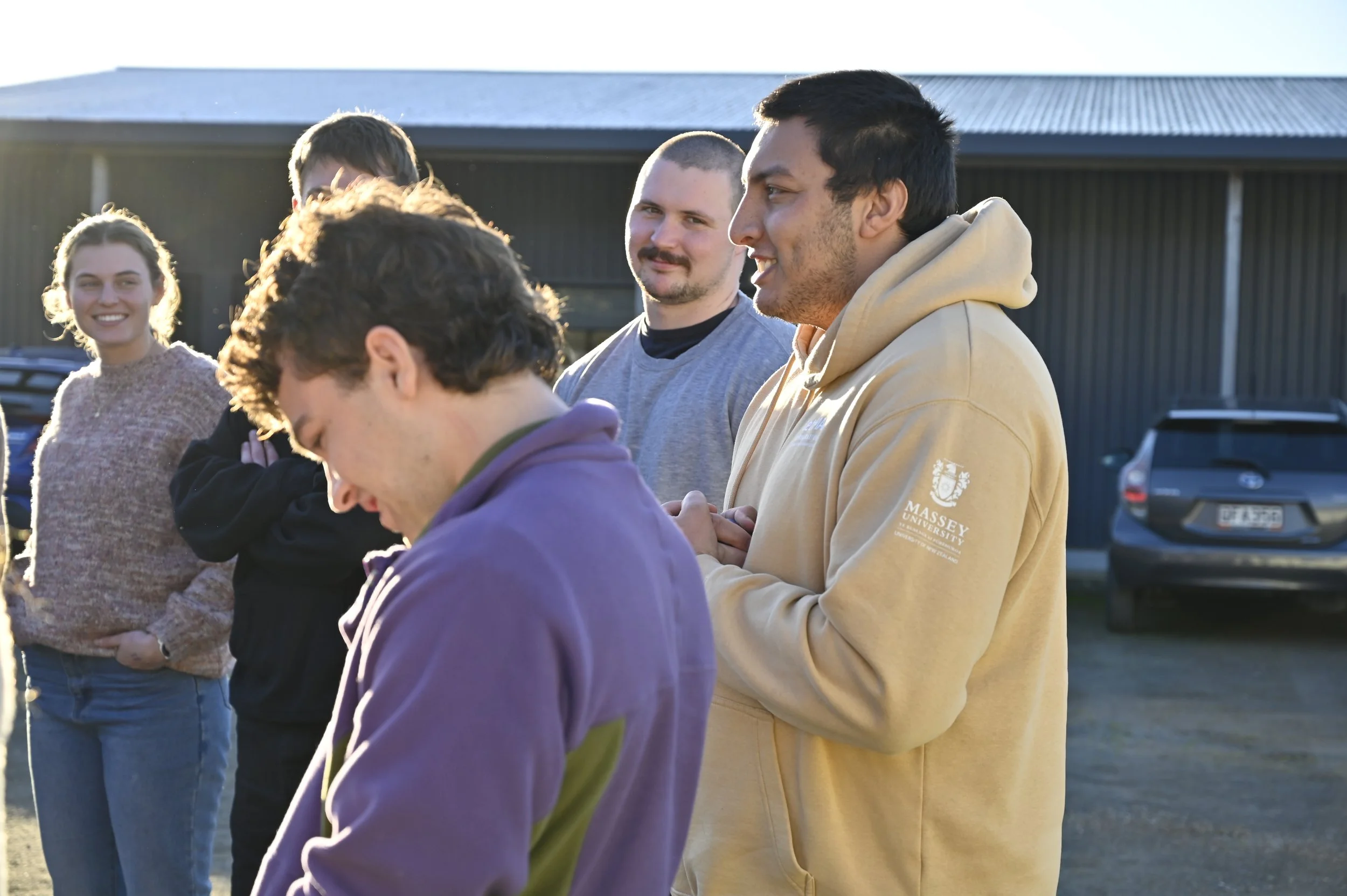 Group of young adults outdoors, smiling and talking, with a building and parked cars in the background.