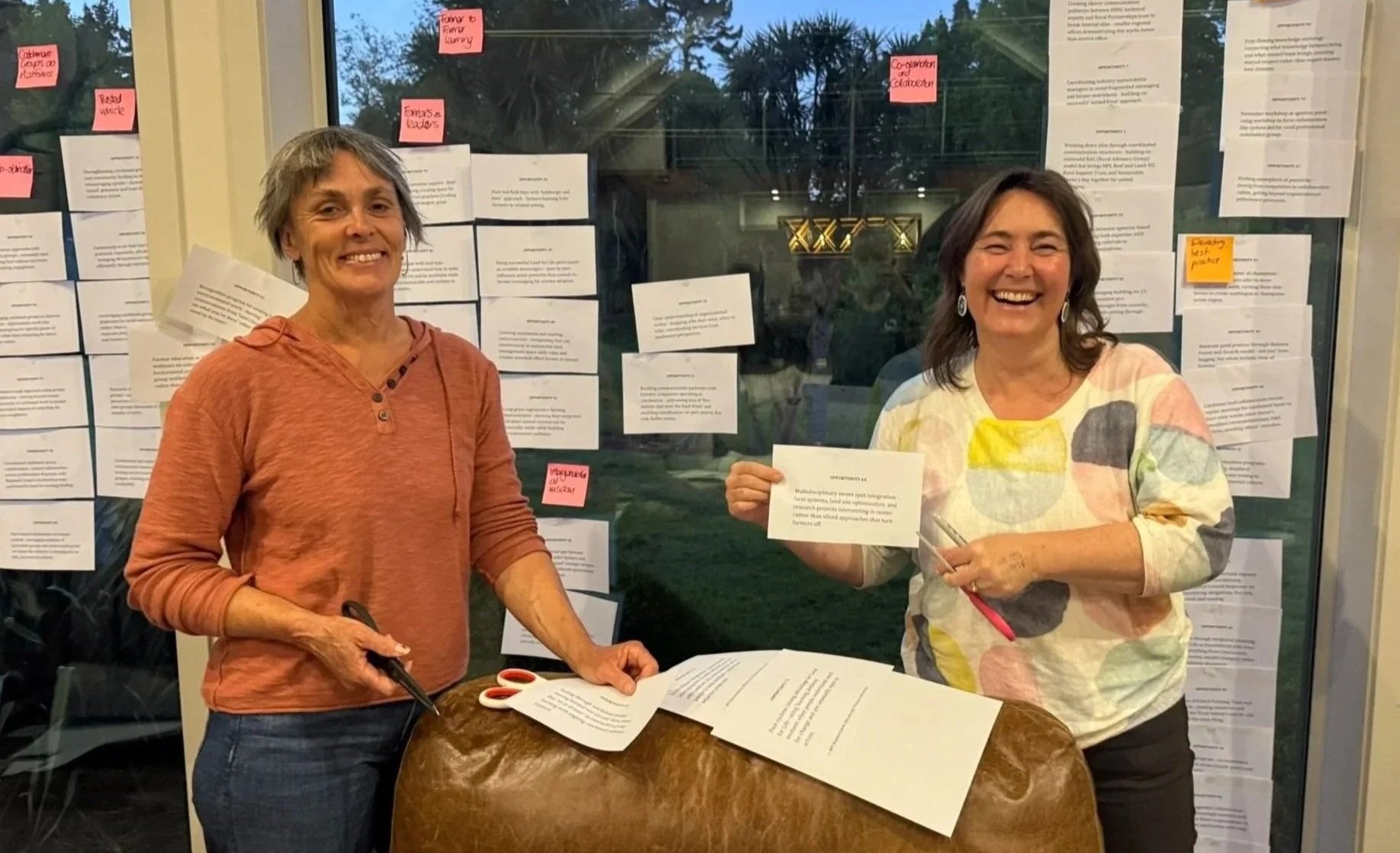 Two women standing at a table smiling, one holding a sheet of paper, with a background of papers and sticky notes on a window.