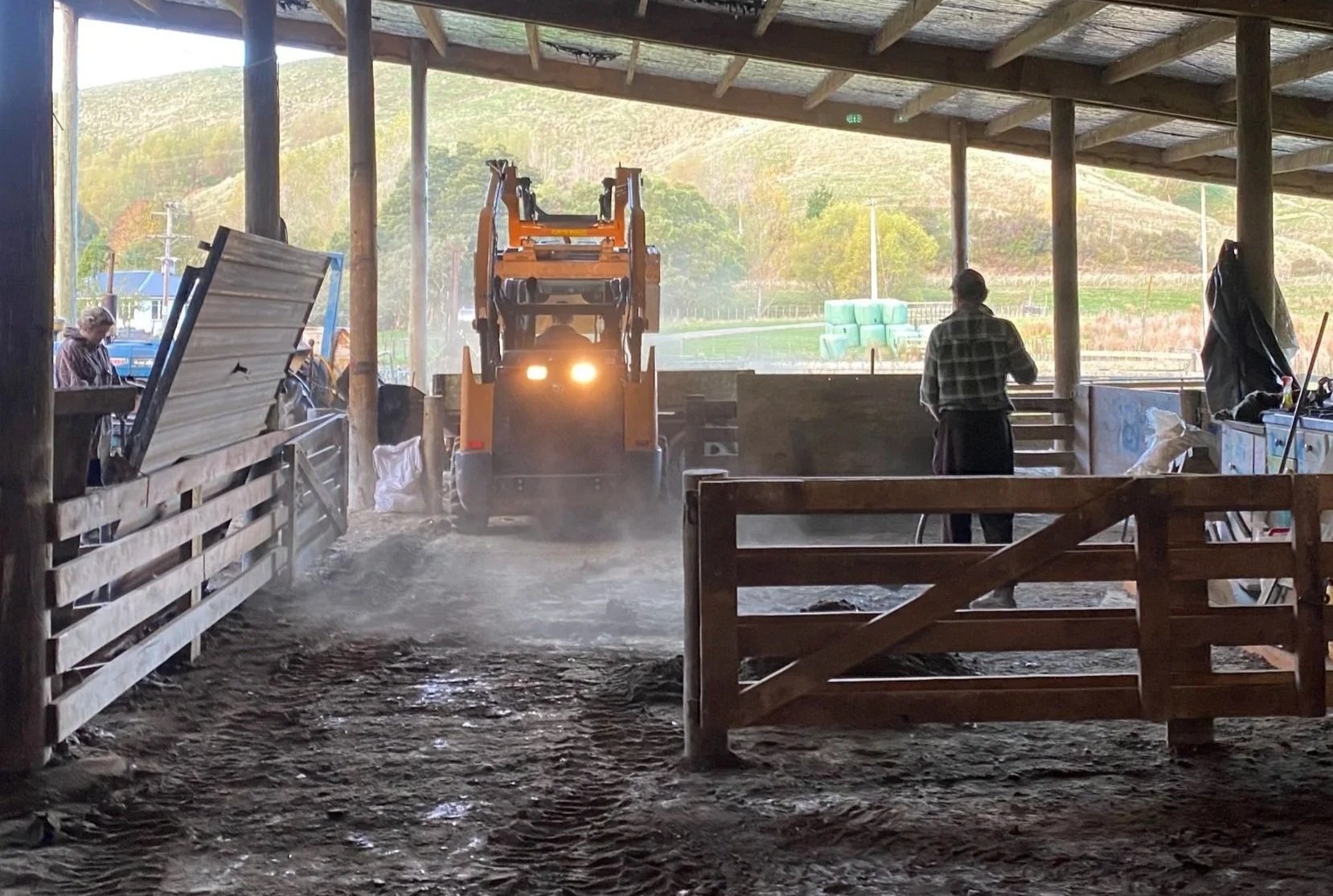 A tractor inside a barn or shed working on the ground with a person nearby observing. There are other people on the left near some equipment, and the barn is open to a field with green hills and hay bales in the background.