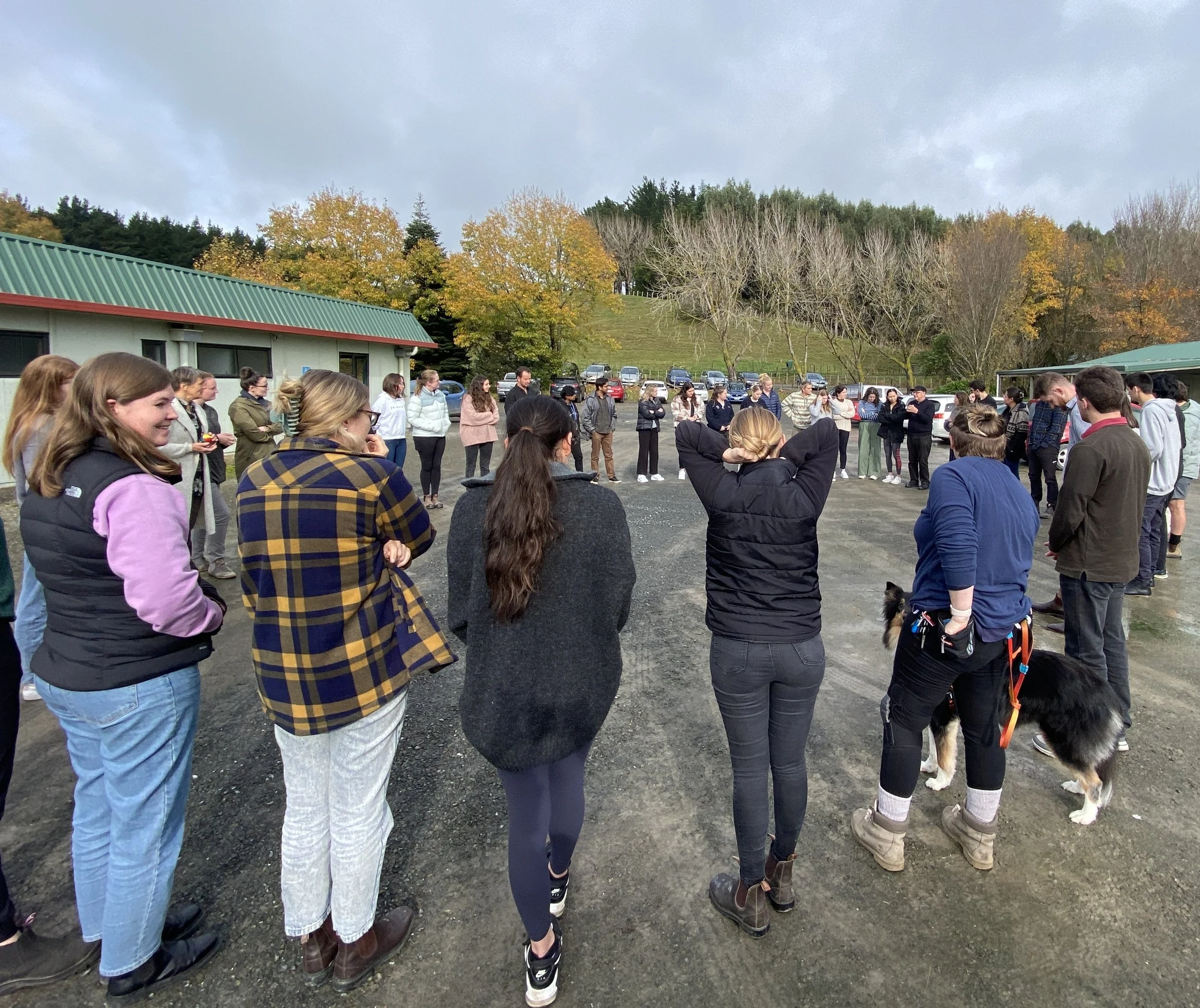 Group of people standing in a circle outdoors on a cloudy day, with trees and parked cars in the background.