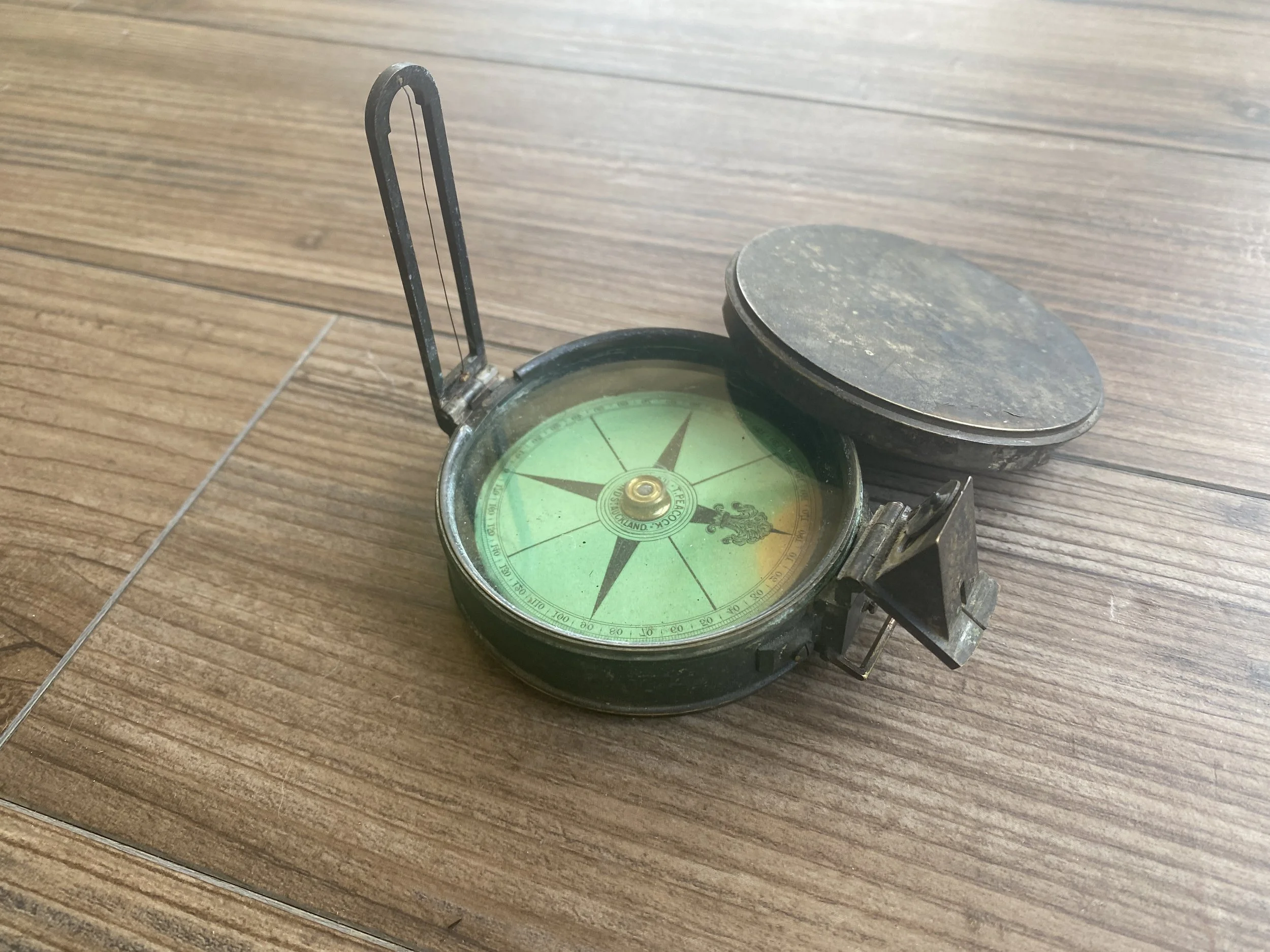 A vintage black compass with a green face and a detailed dial, resting on a wooden surface with a slightly open lid.