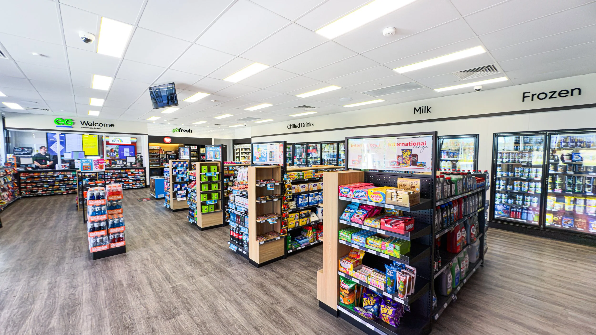 Interior of a convenience store with shelves stocked with snacks, beverages, and frozen goods, a checkout counter, and signage indicating sections like 'Chilled Drinks', 'Milk', and 'Frozen'.