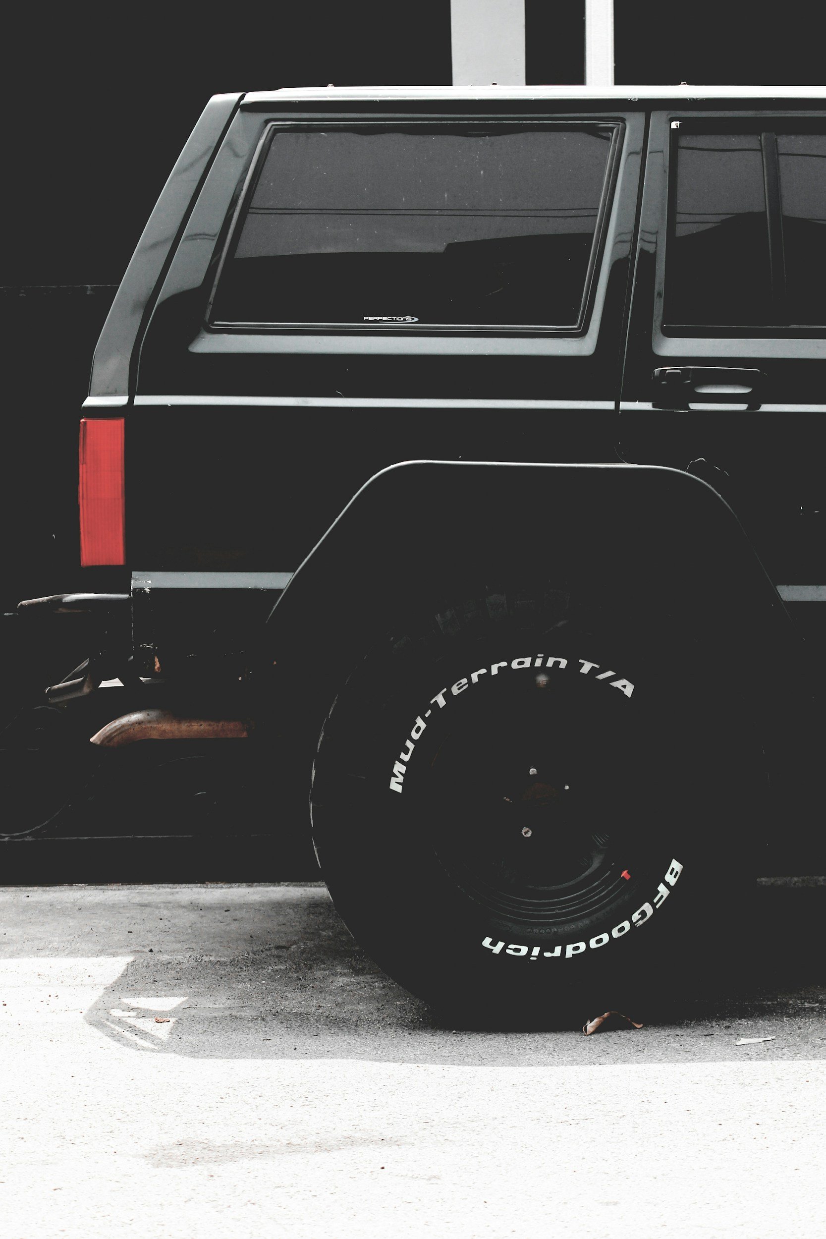 Close-up of the rear side of a black off-road vehicle with a large SVT tire marked 'MUD-TERRAIN T/A' by BFGoodrich, parked next to a white building.