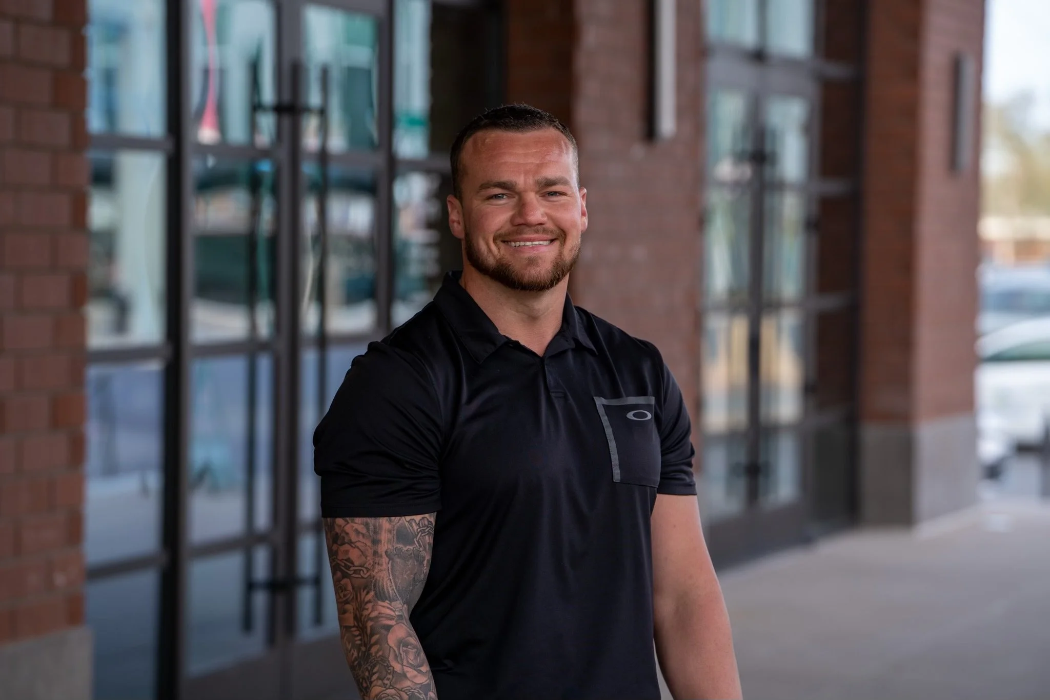 A smiling man with a beard and short hair standing outdoors near a brick building with large windows, wearing a black polo shirt.