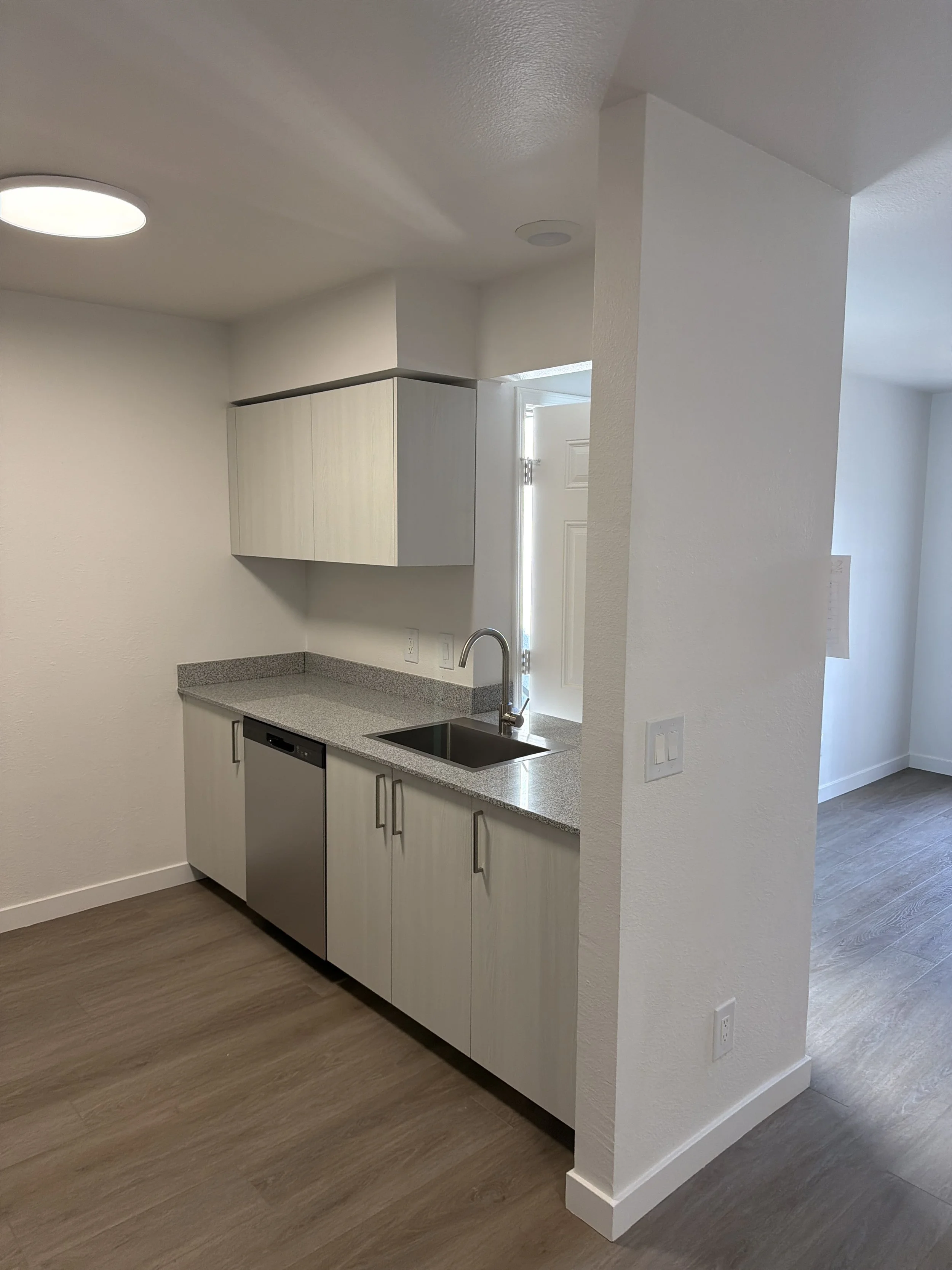 Small kitchen with light-colored cabinets, granite countertop, single sink, and dishwasher, with hardwood flooring and white walls.