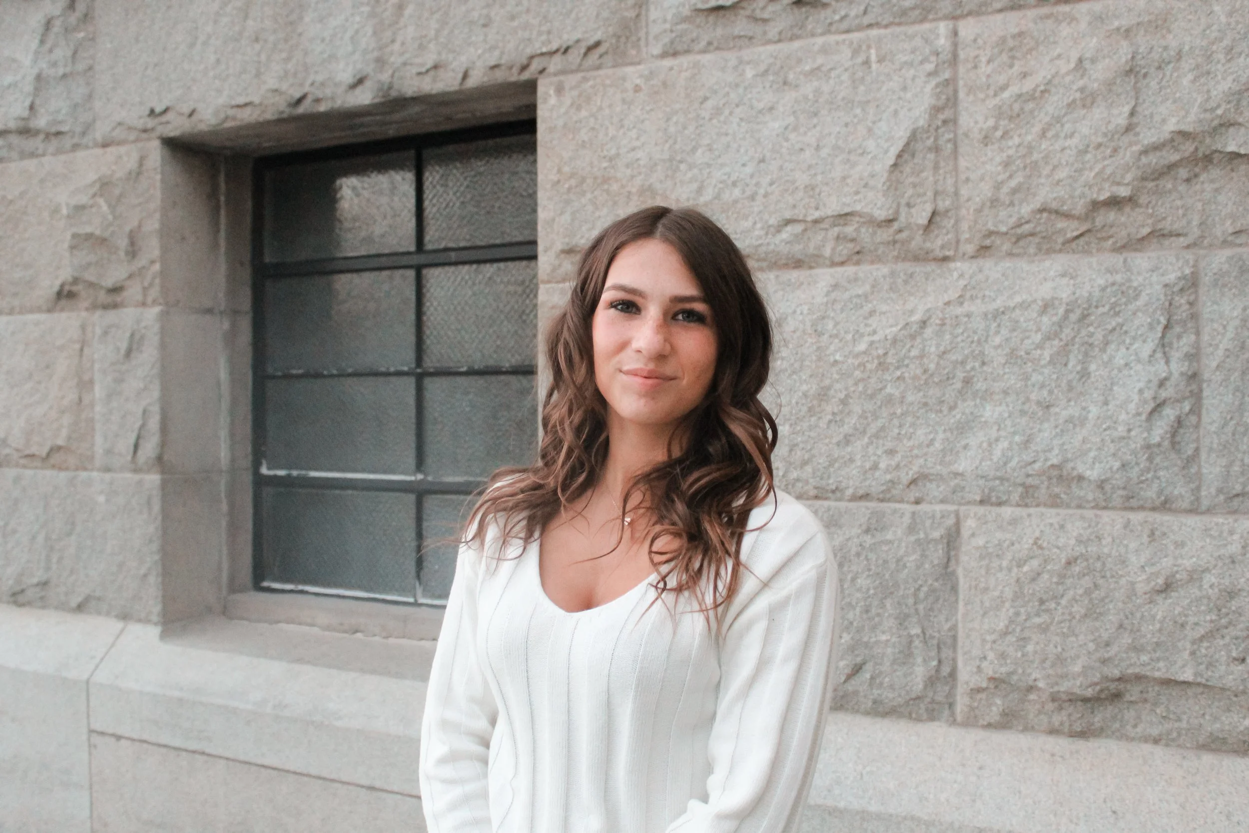 A young woman with wavy brown hair and a light complexion, wearing a white sweater, standing outside against a stone wall with a window.