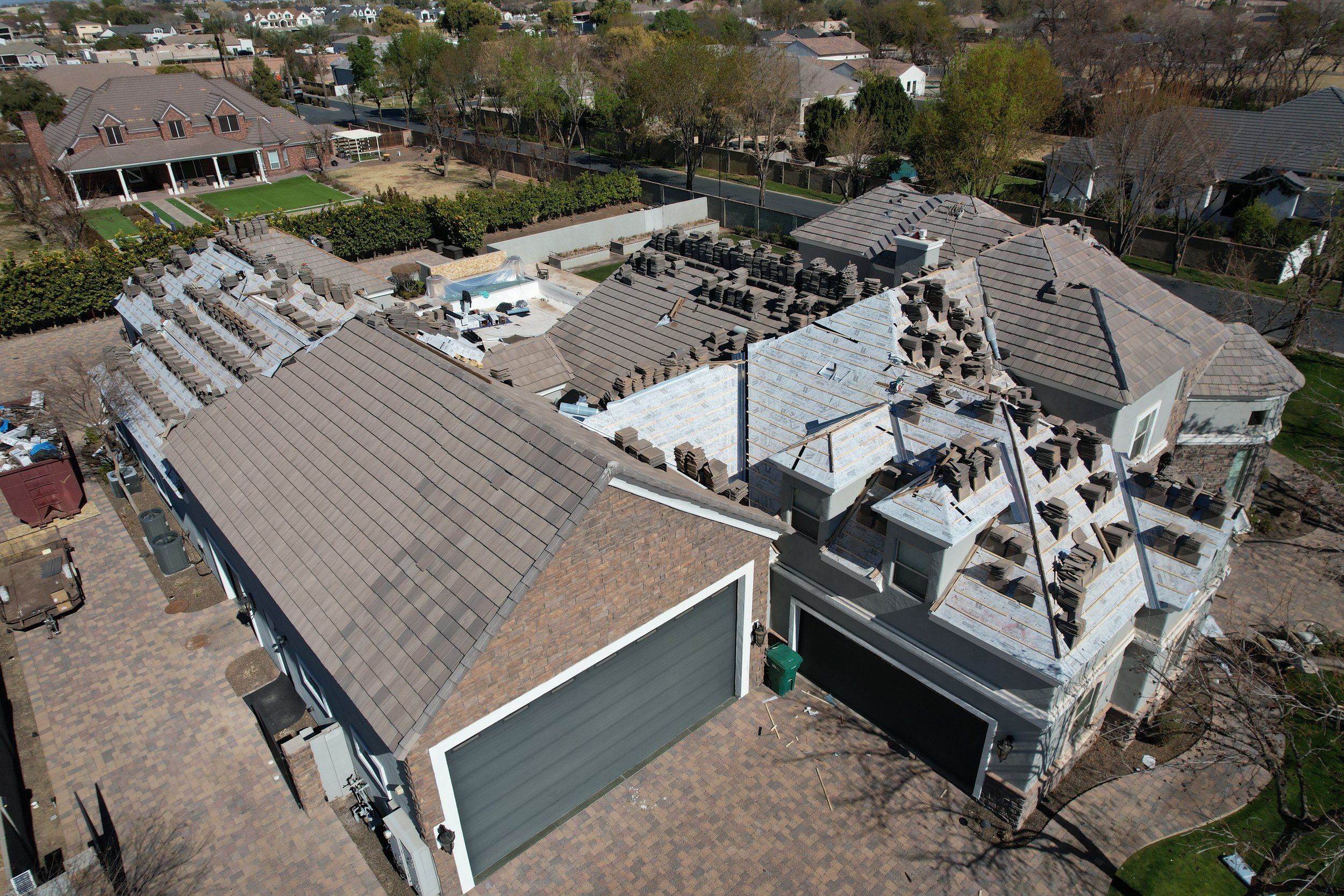 A large house under renovation with scaffolding and roofing materials on the roof, surrounded by trees, neighboring houses, and a paved driveway. Re- Roof