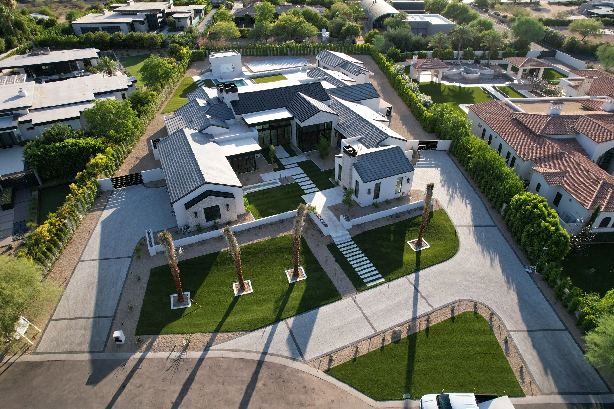 A large modern house in a suburban neighborhood with a manicured lawn, palm trees, and surrounding greenery. The house features white walls, a variety of roof styles, and a paved driveway with a white vehicle parked at the entrance.