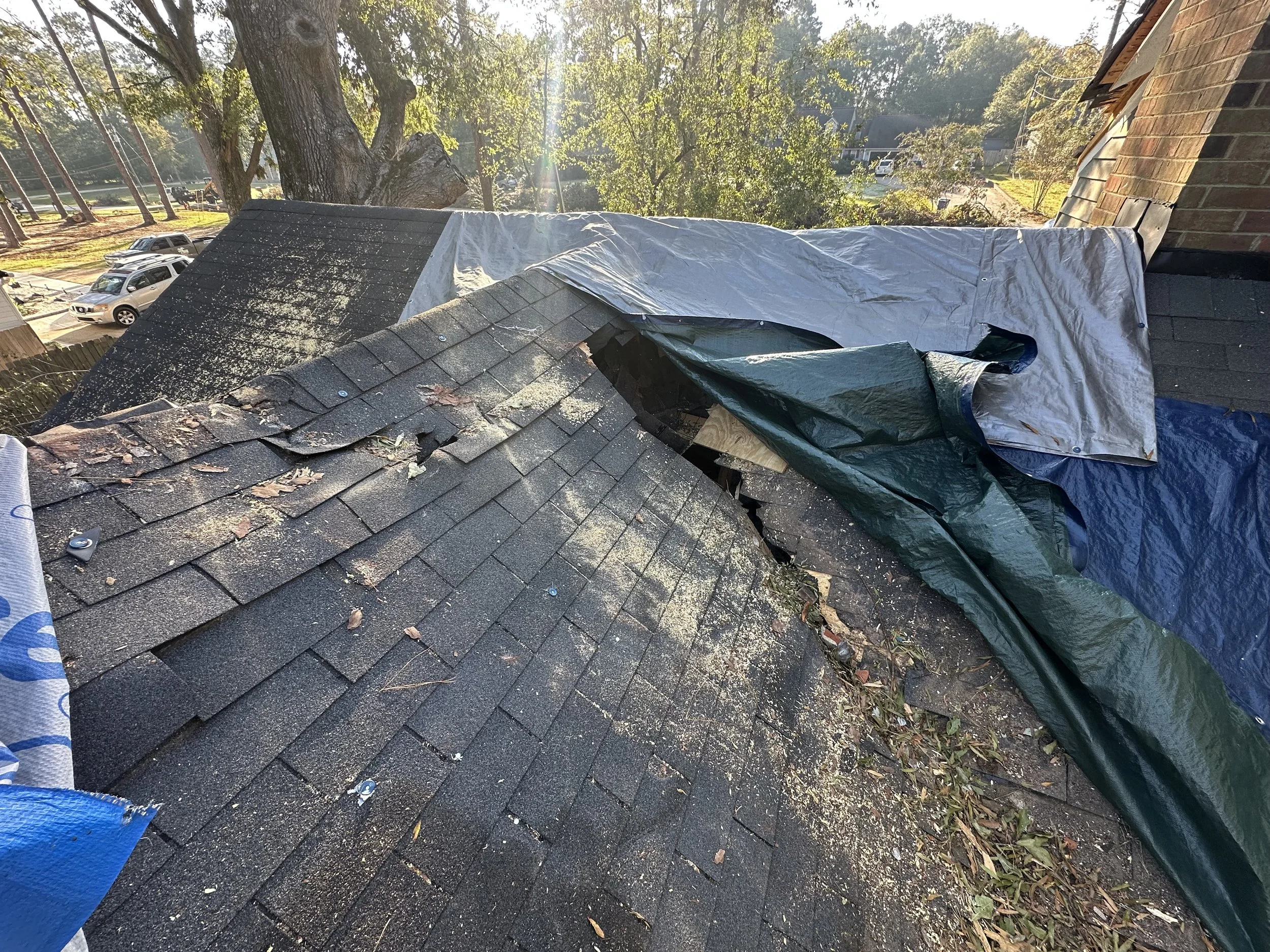 Damaged roof with loose shingles and tarps covering part of the roof, debris scattered on the shingles, and outdoor trees and parked cars in the background.