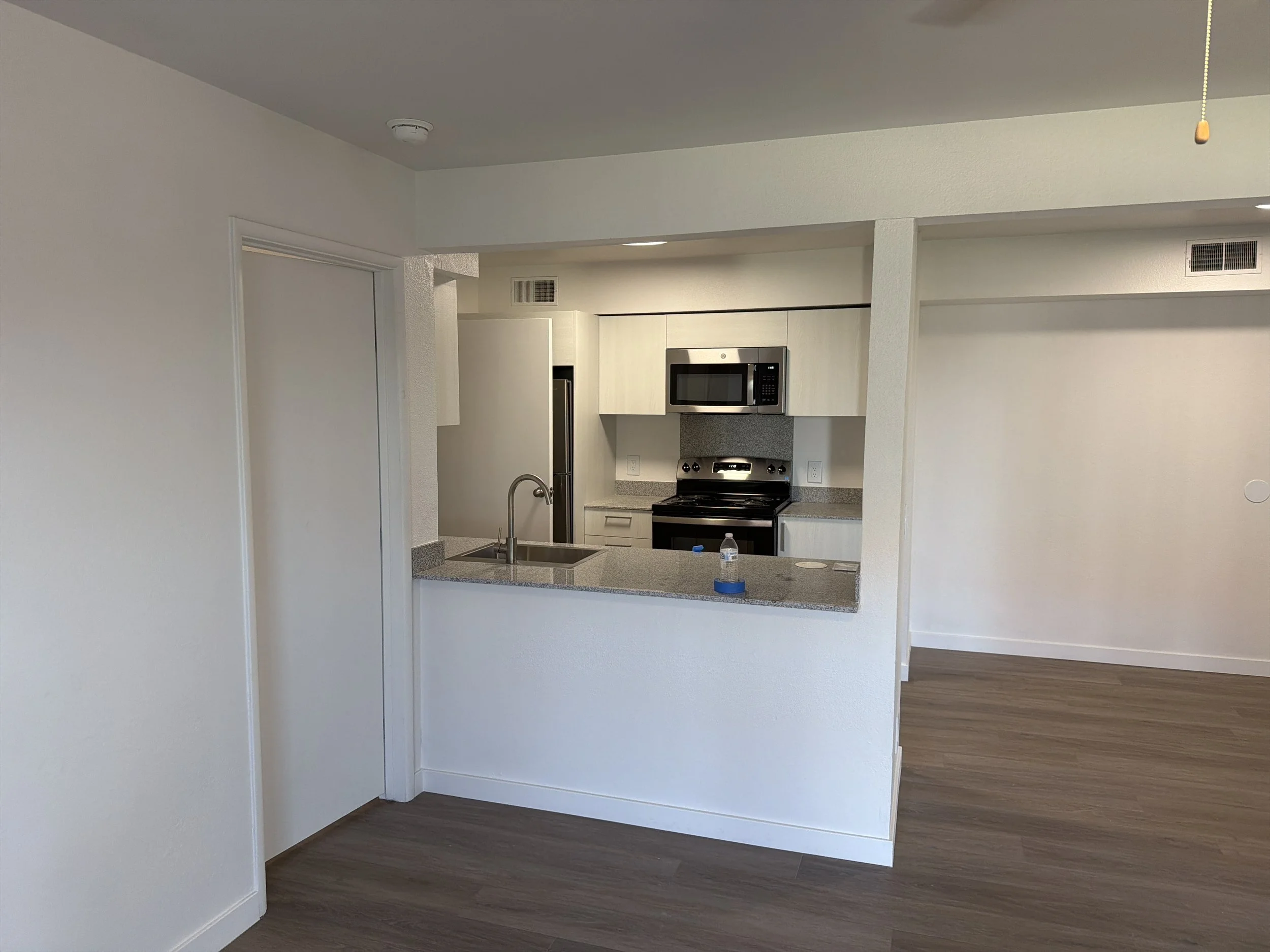 Empty kitchen with white cabinets, stainless steel stove, microwave, and refrigerator, gray granite countertops, and wood flooring.