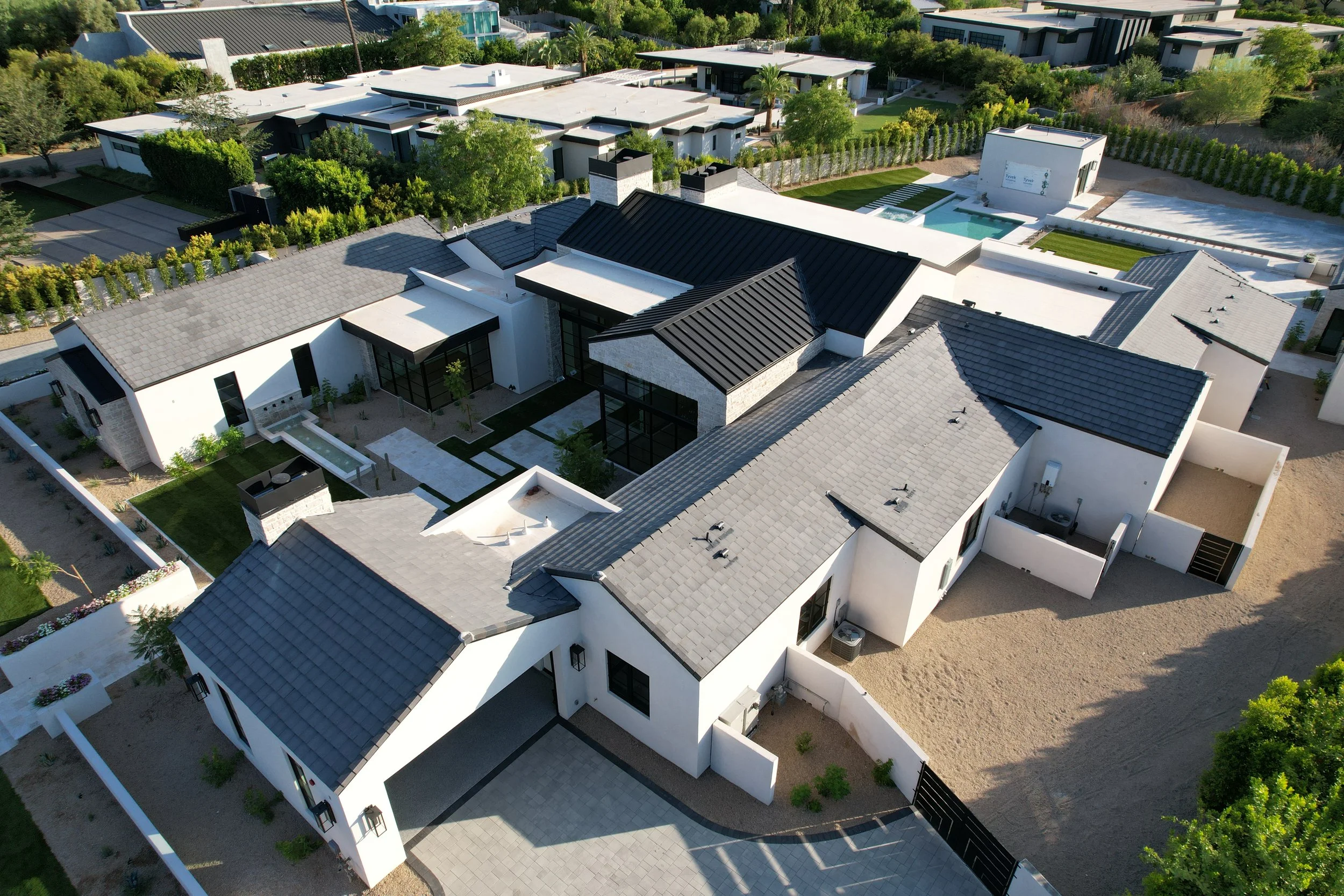 Aerial view of a modern, multi-section house with gray and black roofs, white walls, outdoor patio area, swimming pool, and a landscaped yard.