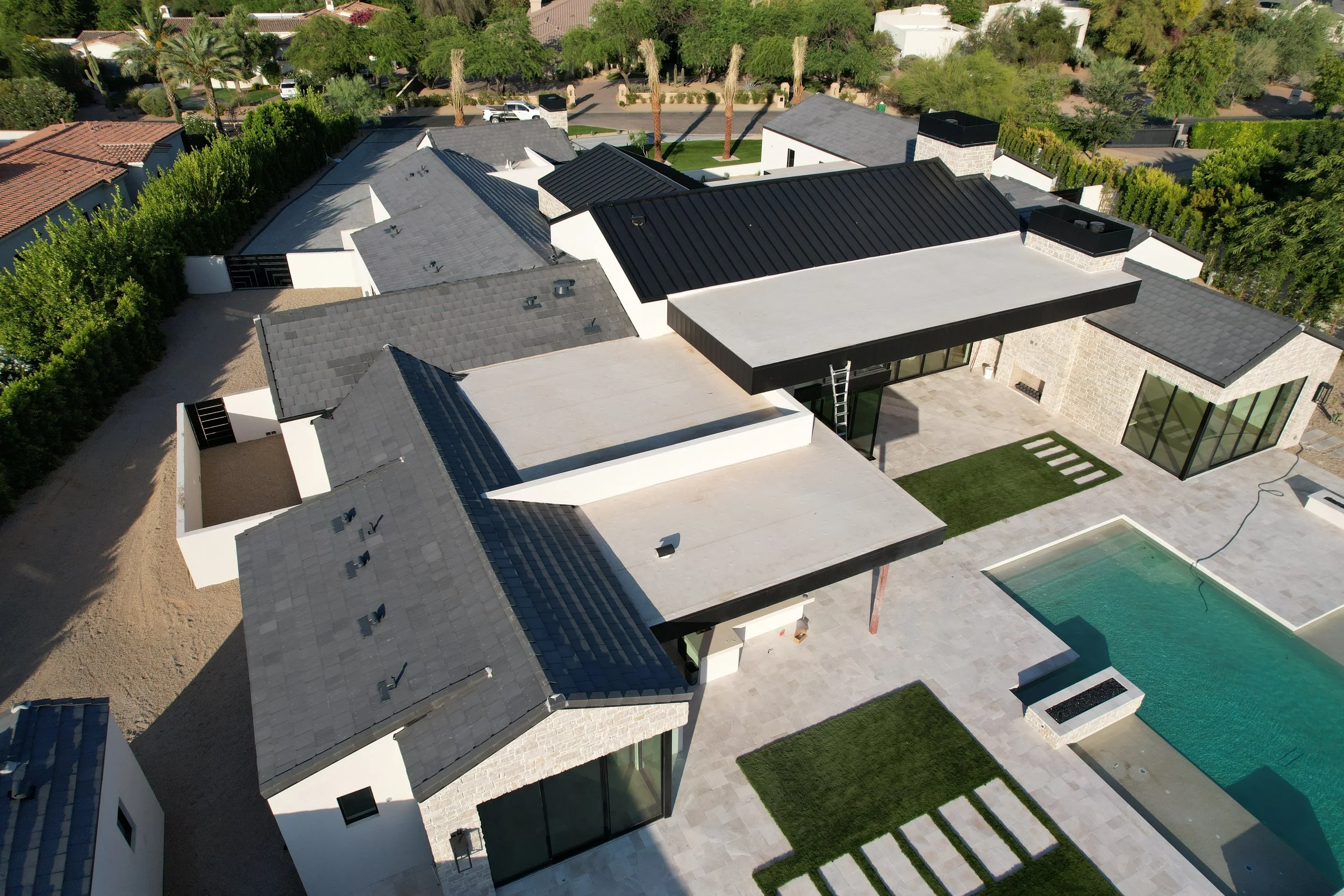 Aerial view of a modern house with a backyard featuring a swimming pool, patio area, and landscaped grass sections, surrounded by trees and neighboring houses. Tile Roof system, TPO roof System