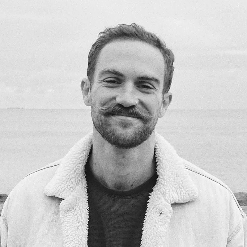 Aiden Canter with a moustache smiling outdoors, wearing a jacket with a shearling collar, in black and white.