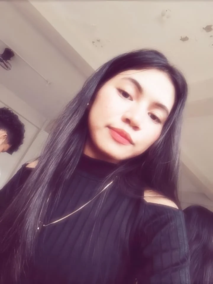 A young woman with long dark hair wearing a black top and a silver necklace, taking a selfie in an indoor setting with part of her face slightly tilted.