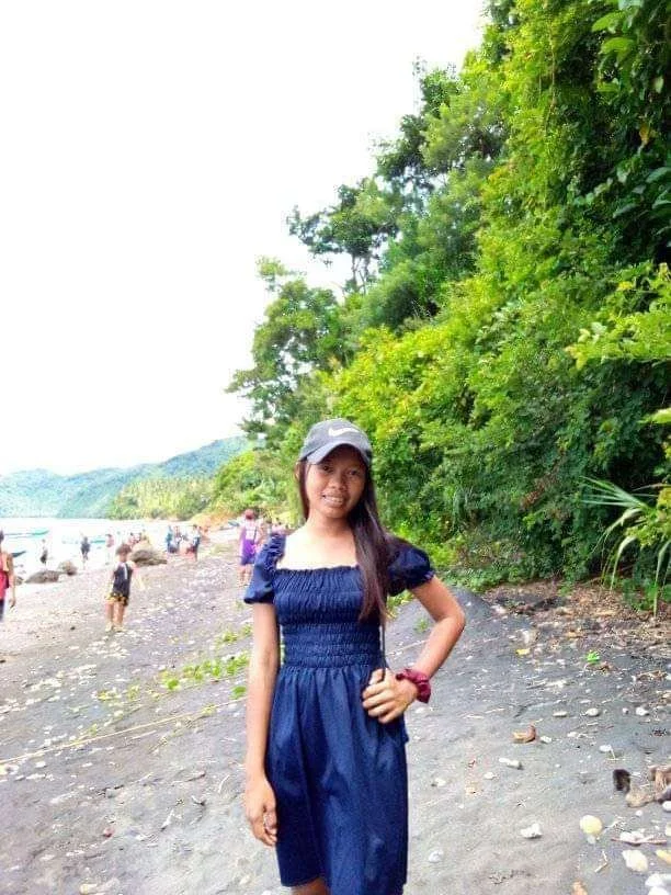 Young woman in a navy blue dress and gray Nike cap standing on a beach with green trees in the background. Other people are visible in the distance on the beach.