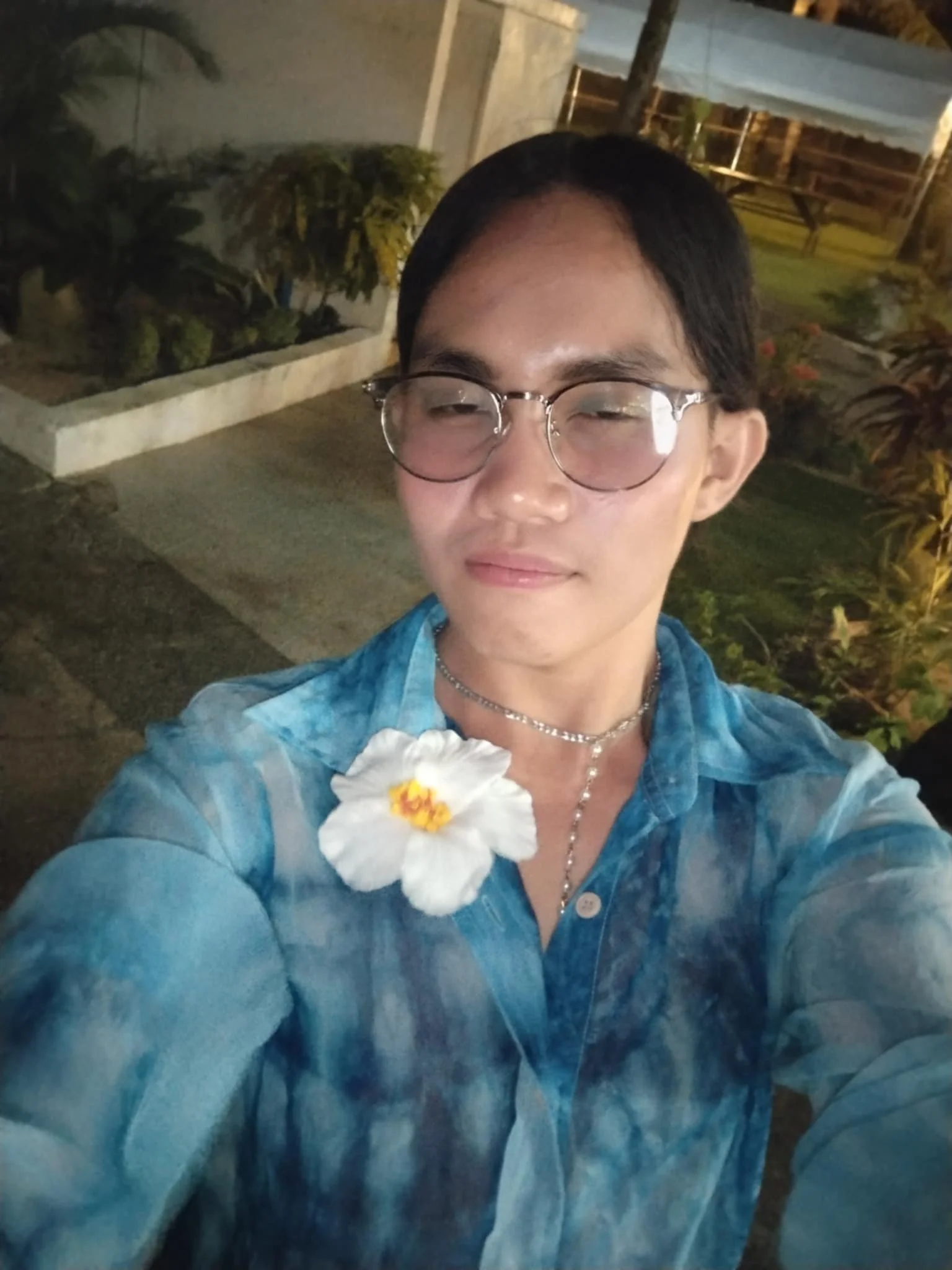 A person with glasses and dark hair is taking a selfie outdoors at night, wearing a blue patterned shirt with a white flower pinned to it, and a necklace.