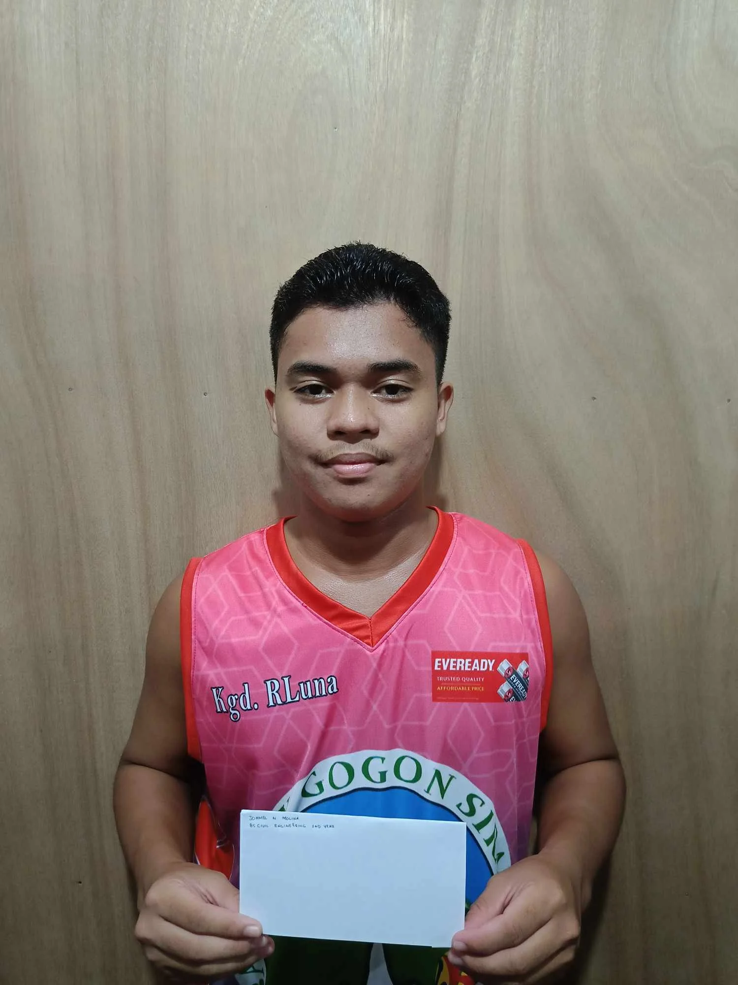 A young man in a pink basketball jersey holding a white envelope stands against a wooden background.