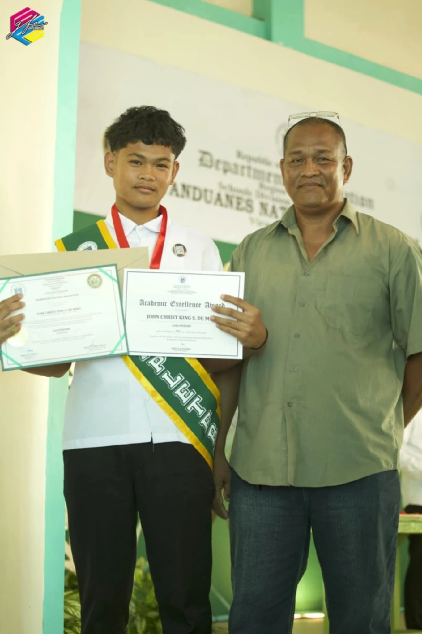 A young student in a school uniform with a sash and medal holding a certificate of academic achievement, standing next to an older man in a green shirt, both smiling, at an awards ceremony.