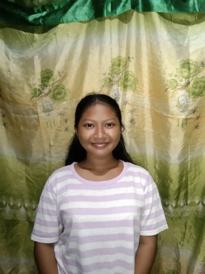 A young woman with medium skin tone, black hair, and a striped pink and white T-shirt standing in front of a decorated curtain with green, yellow, and floral patterns.