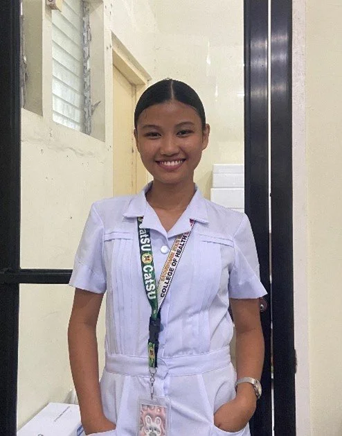 A young girl in a school uniform standing indoors with a smile, wearing a lanyard with an ID badge.