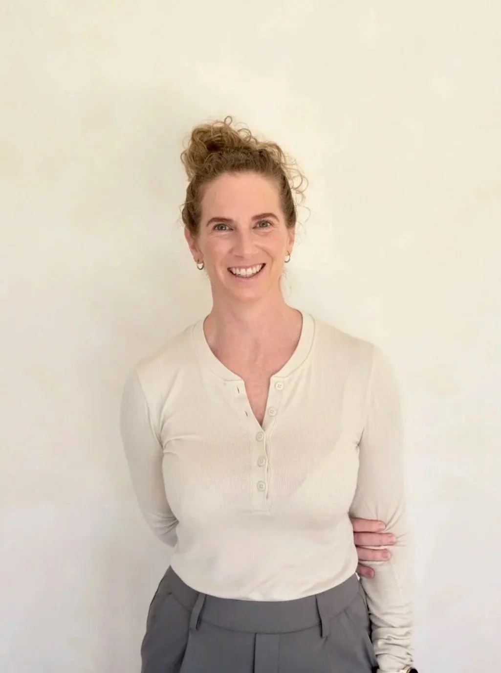 A woman with curly red hair in a bun, smiling, stands against a plain white wall, wearing a beige long-sleeved button-up top and gray pants.