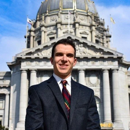 A man in a suit standing in front of a historic government building with a dome and columns.