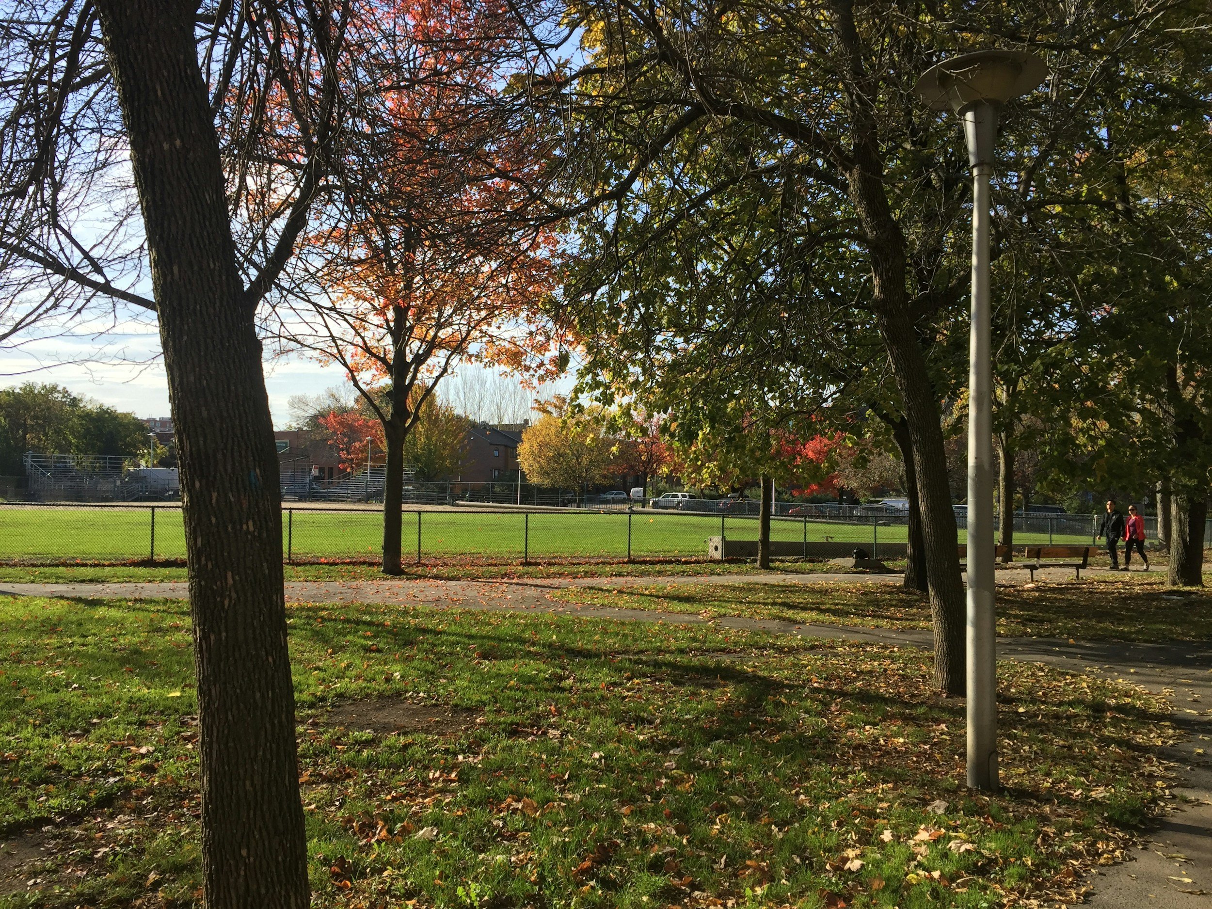 A park with trees, a grassy area, a walking path, a bench, a fence, and two people walking in the distance on a sunny autumn day.