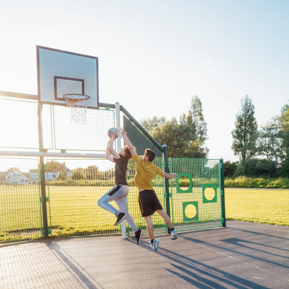 Two boys playing basketball outdoors near a basketball hoop on a sunny day with green trees in the background.