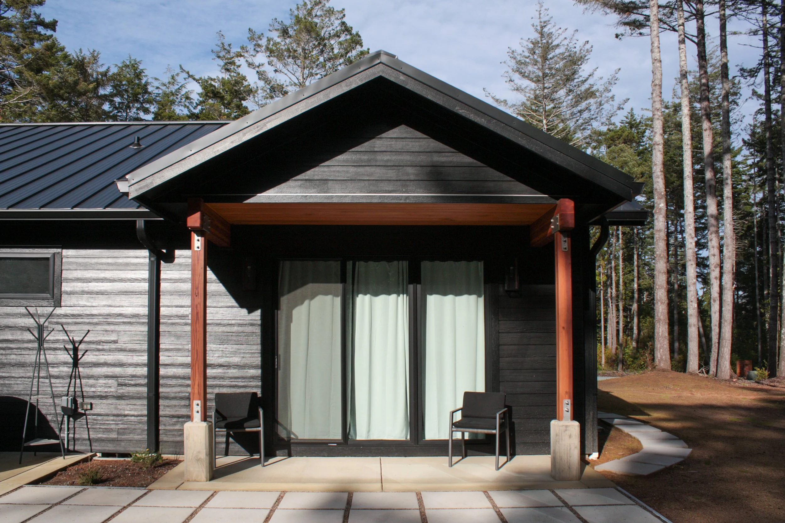 Modern house with black siding, sliding glass door with white curtains, covered porch with two black chairs, and surrounding trees.