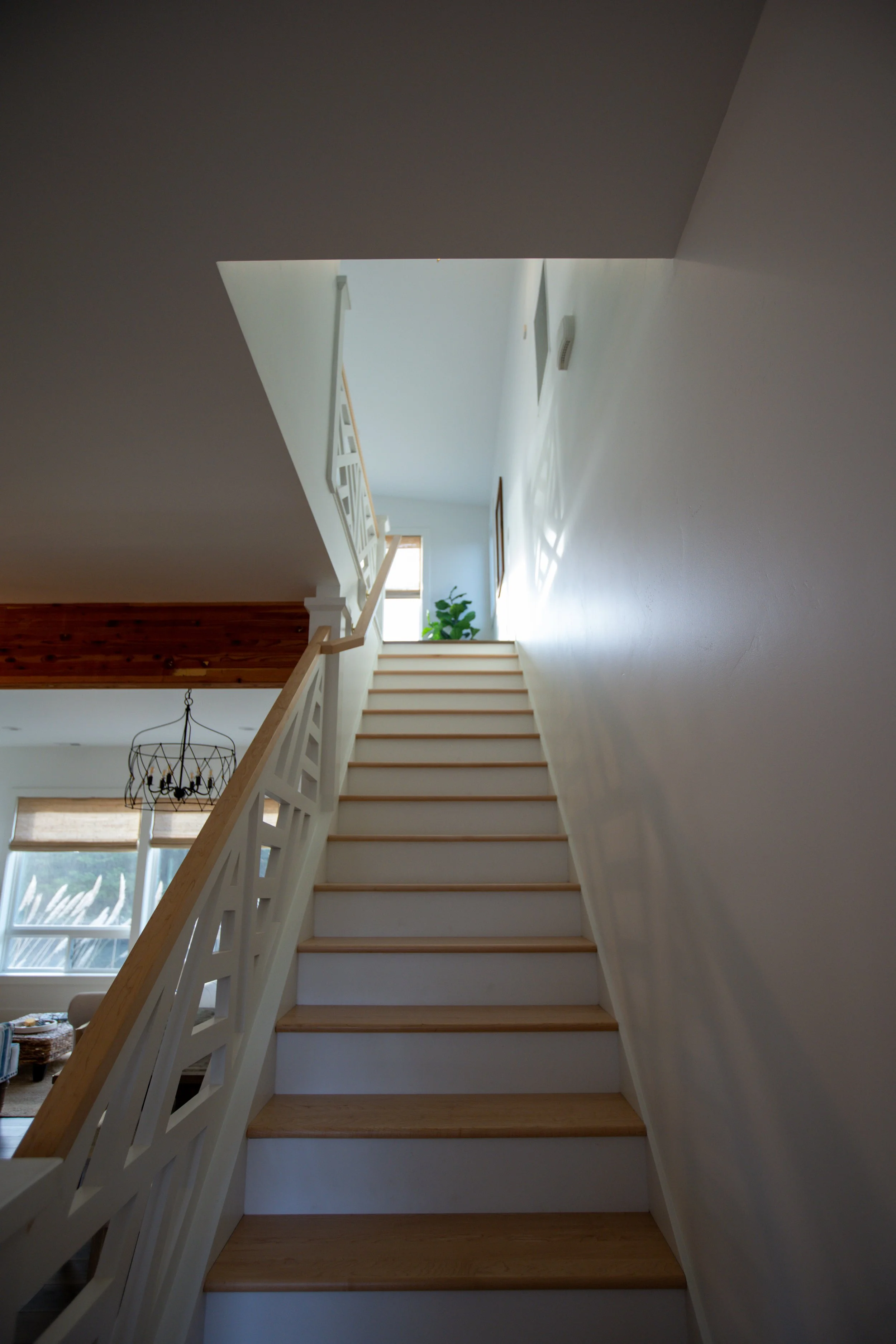 Interior view of a staircase with wooden steps and white railing, leading up to a bright landing with a small window, a potted plant, and framed artwork on the wall.