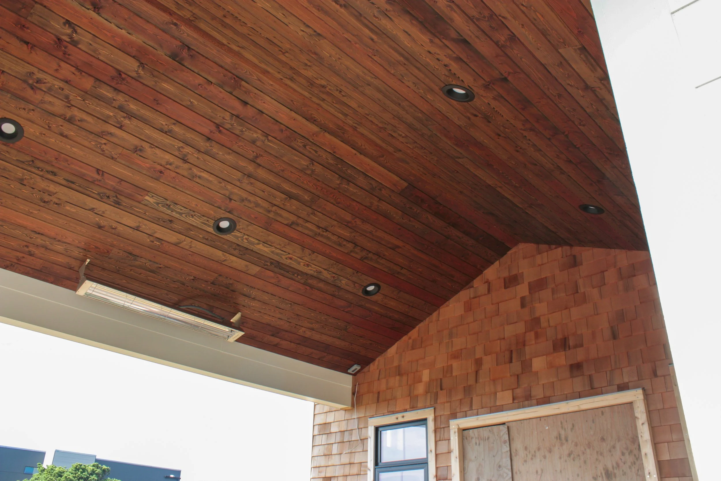 Ceiling of a building with wooden planks and recessed lighting, part of an exterior overhang, with a section of brick wall and a window below.
