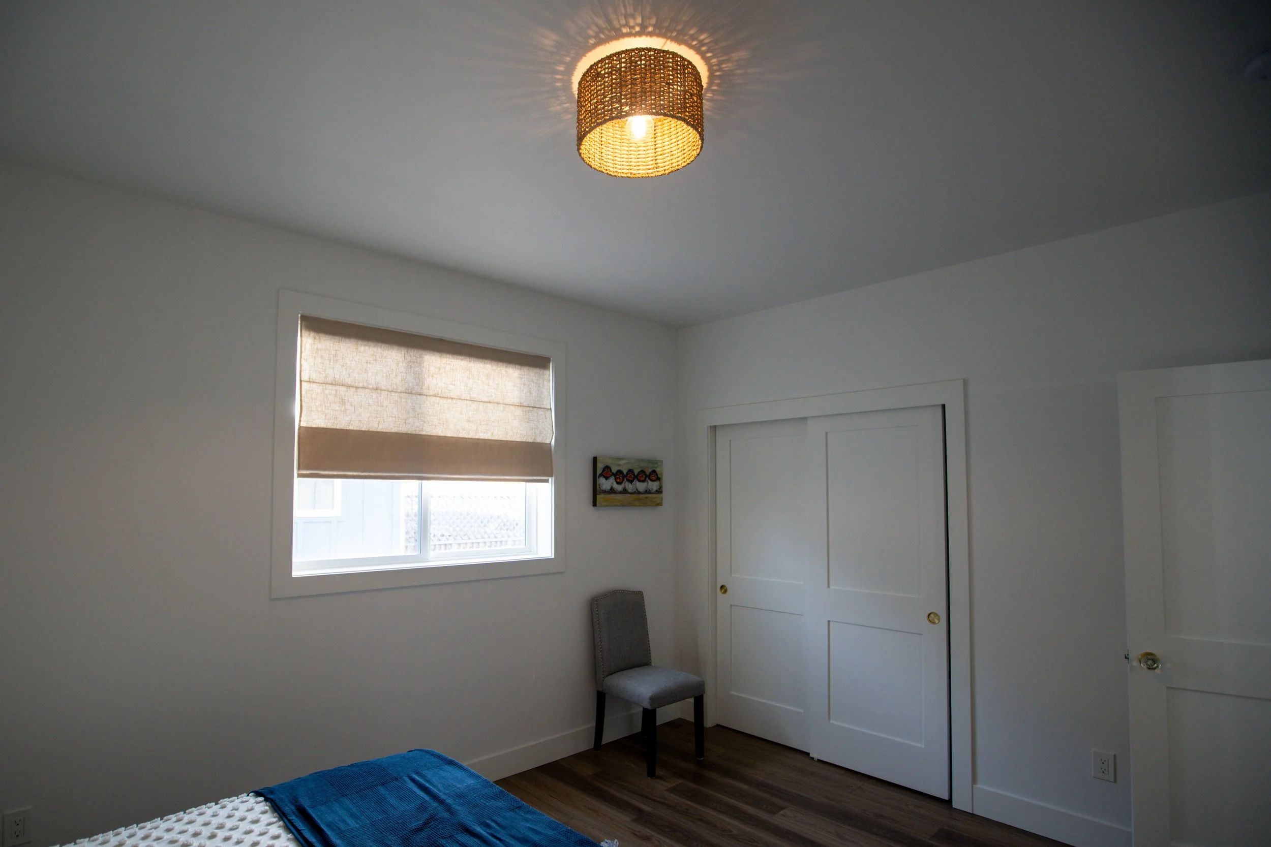 A minimalist bedroom with white walls, a window with a beige Roman shade, a closed white closet door, a gray upholstered chair, a colorful small painting, and a bed with a white blanket and blue throw blanket. The ceiling light has a woven rattan sha