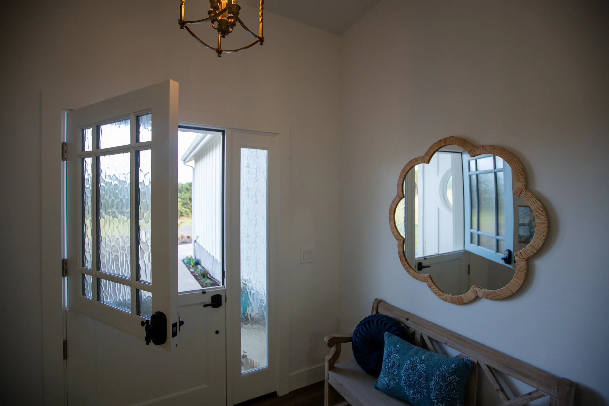 Interior view of a hallway with a white front door with glass panels and a black handle, a decorative mirror on the wall with a wicker frame, a wooden bench with a blue cushion, and a window showing an outdoor view.