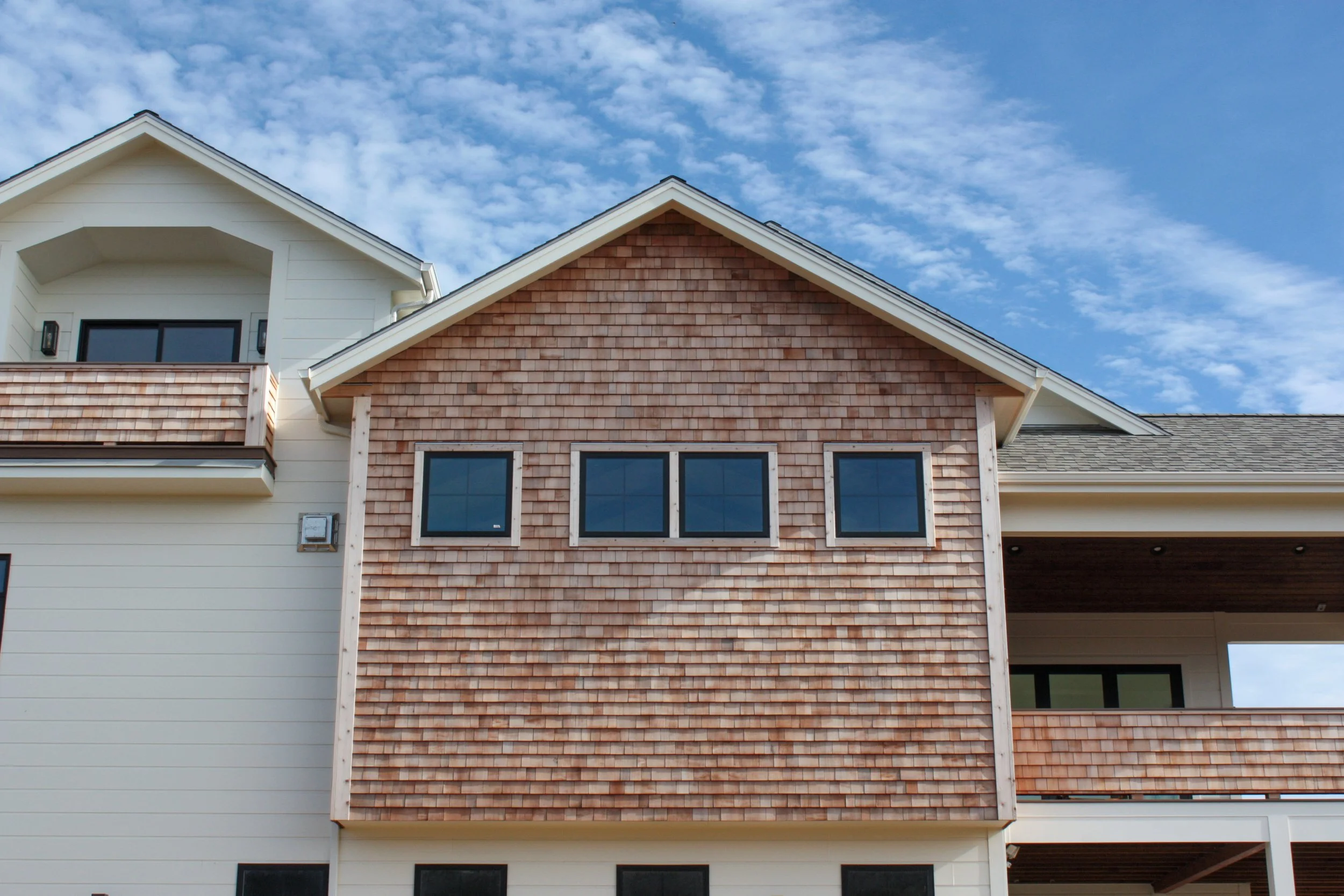 Front view of a modern house with a mix of white siding and reddish-brown shingle siding, four windows on the upper part, and a balcony on the left side, under a blue sky with scattered clouds.