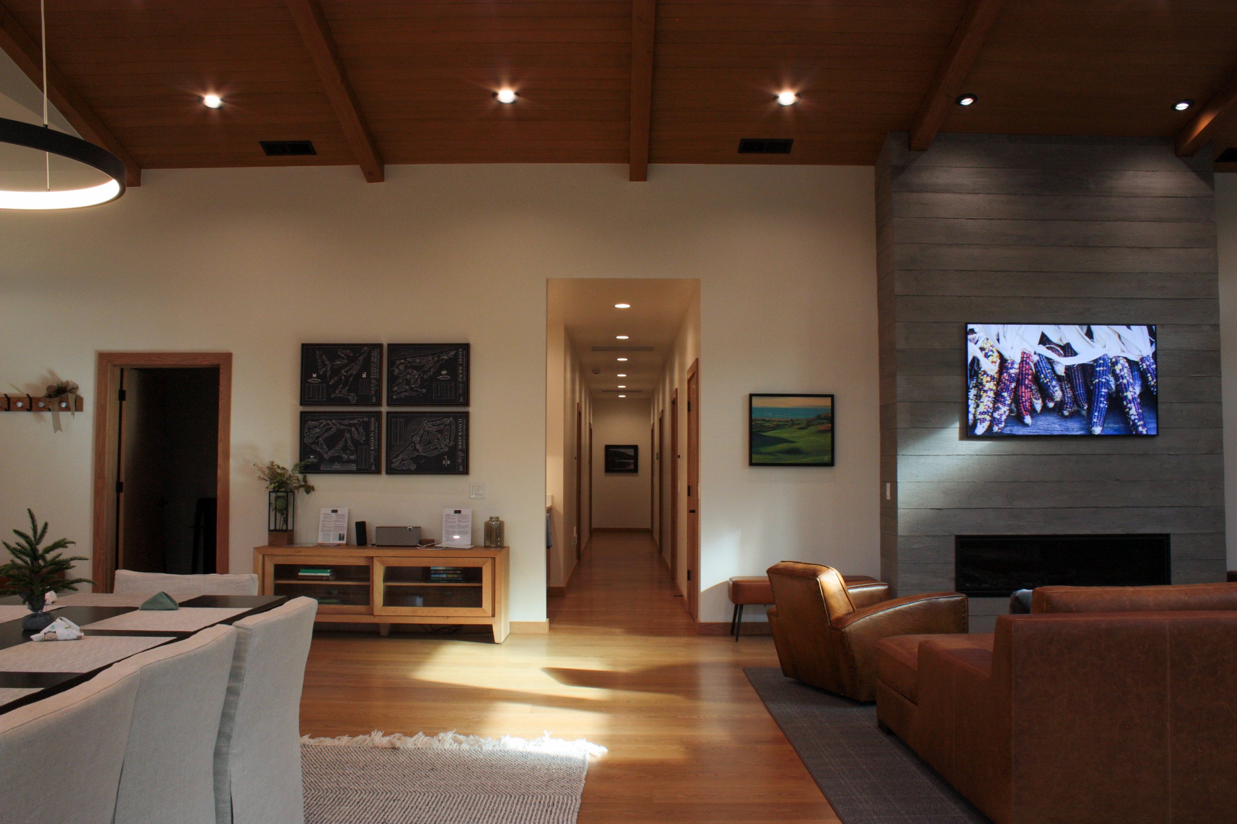 Living room with wooden ceiling and floor, featuring a brown leather armchair, a beige couch, a mounted flat-screen TV with colorful shoes display, and artwork on the walls. A hallway leading to other rooms is visible in the background.