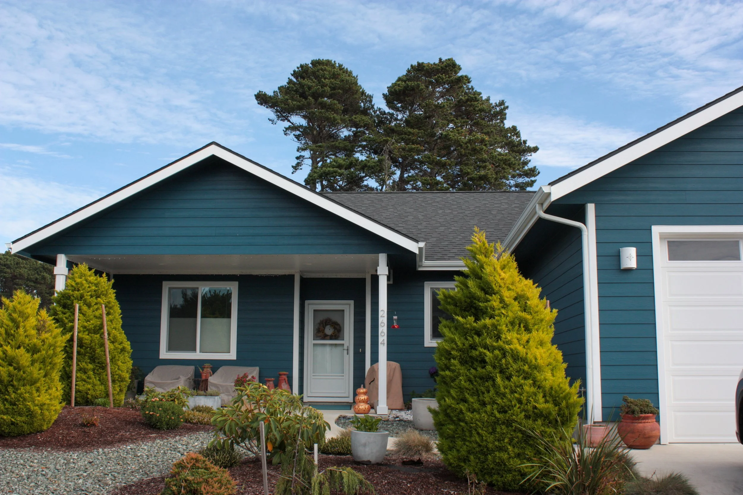 A blue house with a front porch, white trim, and a white garage door. There are green shrubs and potted plants in the front yard, and a clear blue sky overhead.