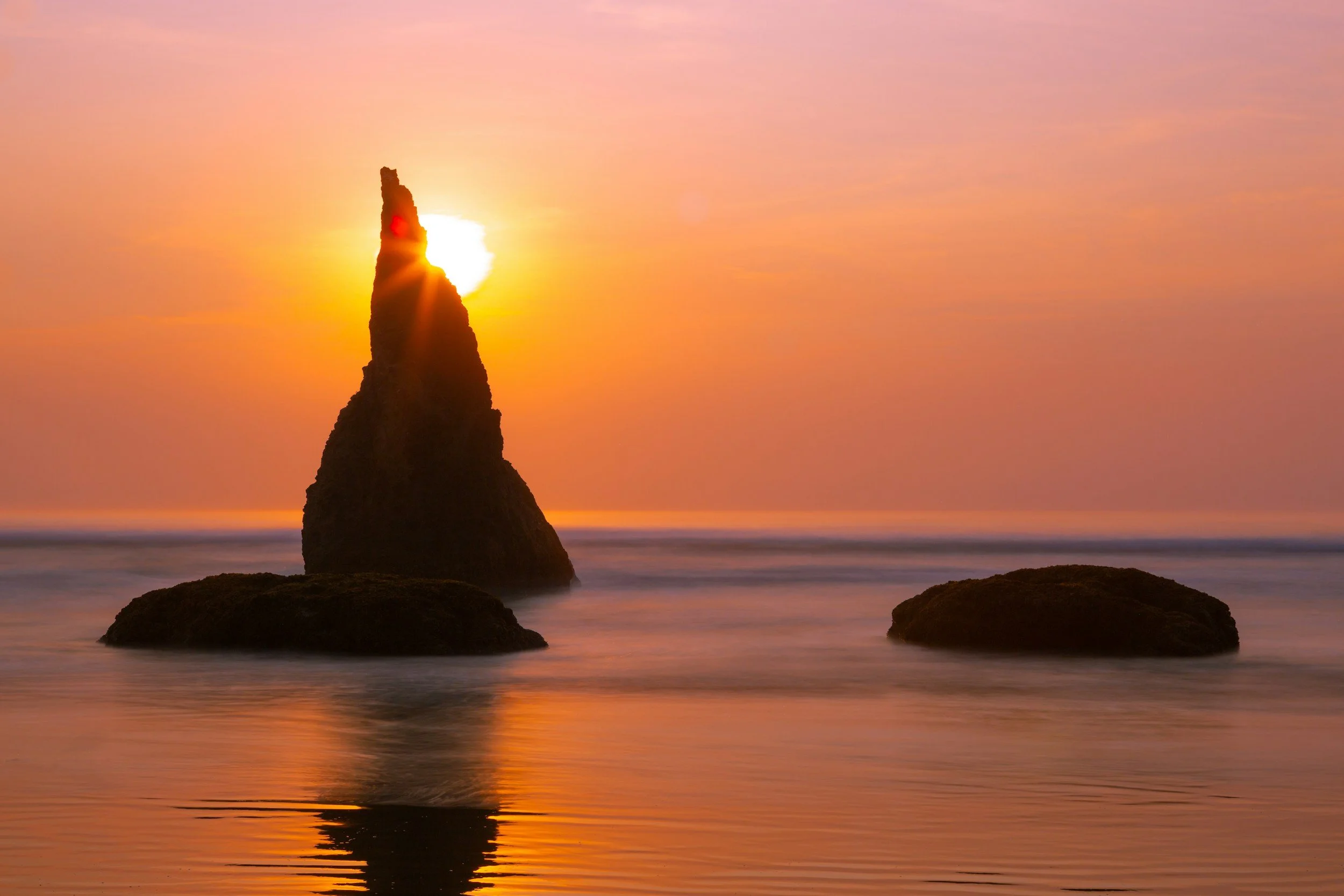 Sunset over the ocean with a tall, jagged rock formation and two smaller rocks in the water, pink and orange sky.