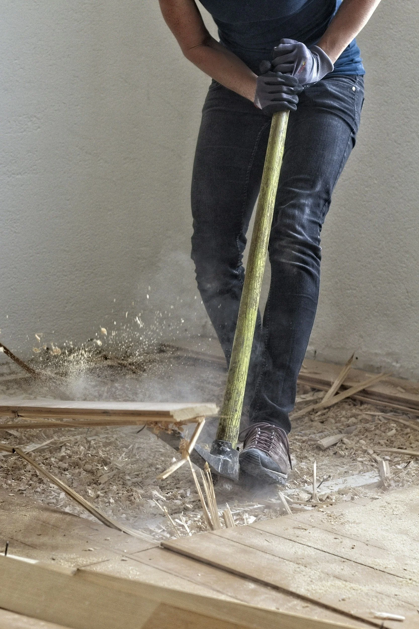 A person wearing gloves and jeans using a sledgehammer to break up wood flooring.