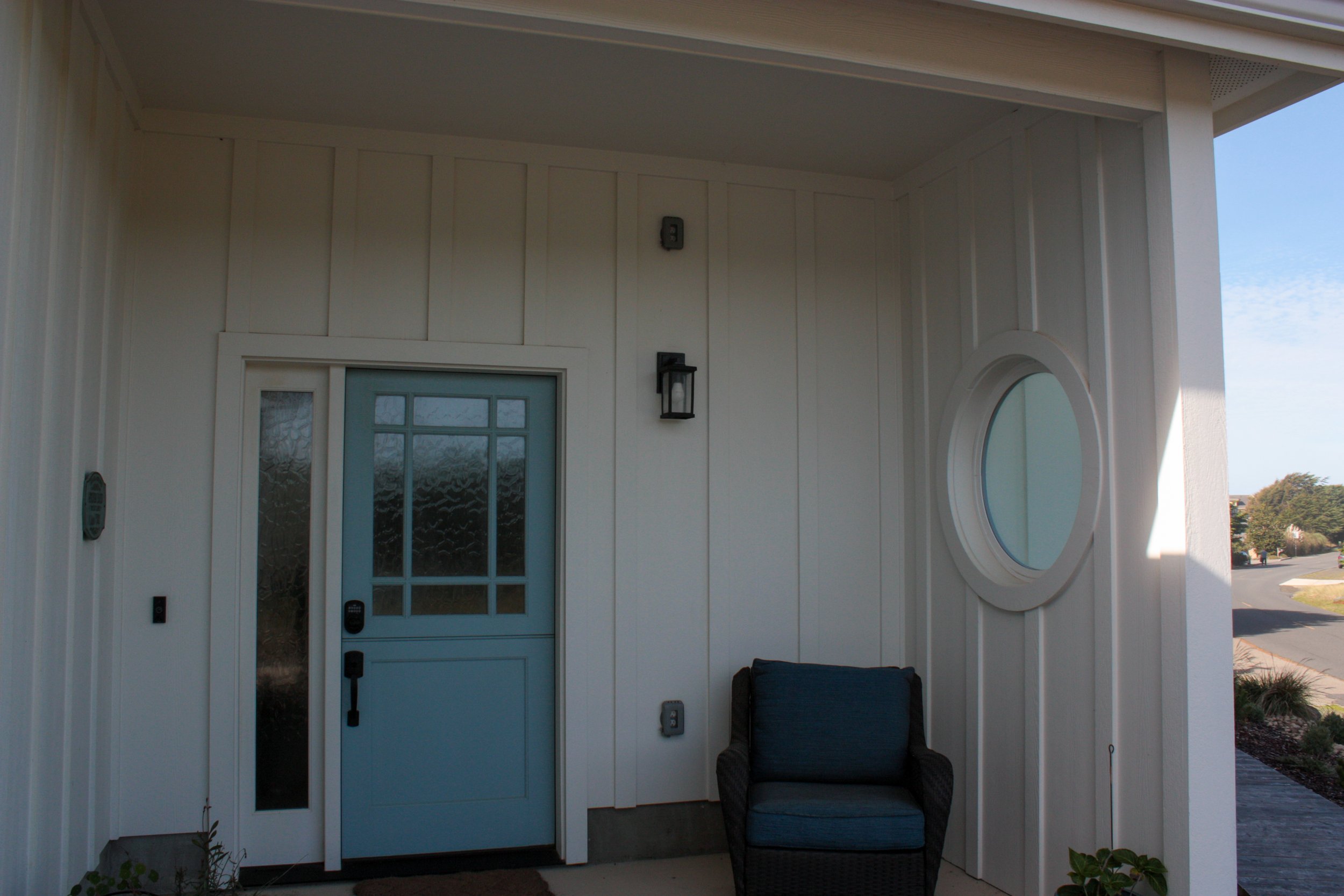 Front porch area of a house with a light blue door, window, black wall-mounted light, and black outdoor chair, with a small plant on right side.