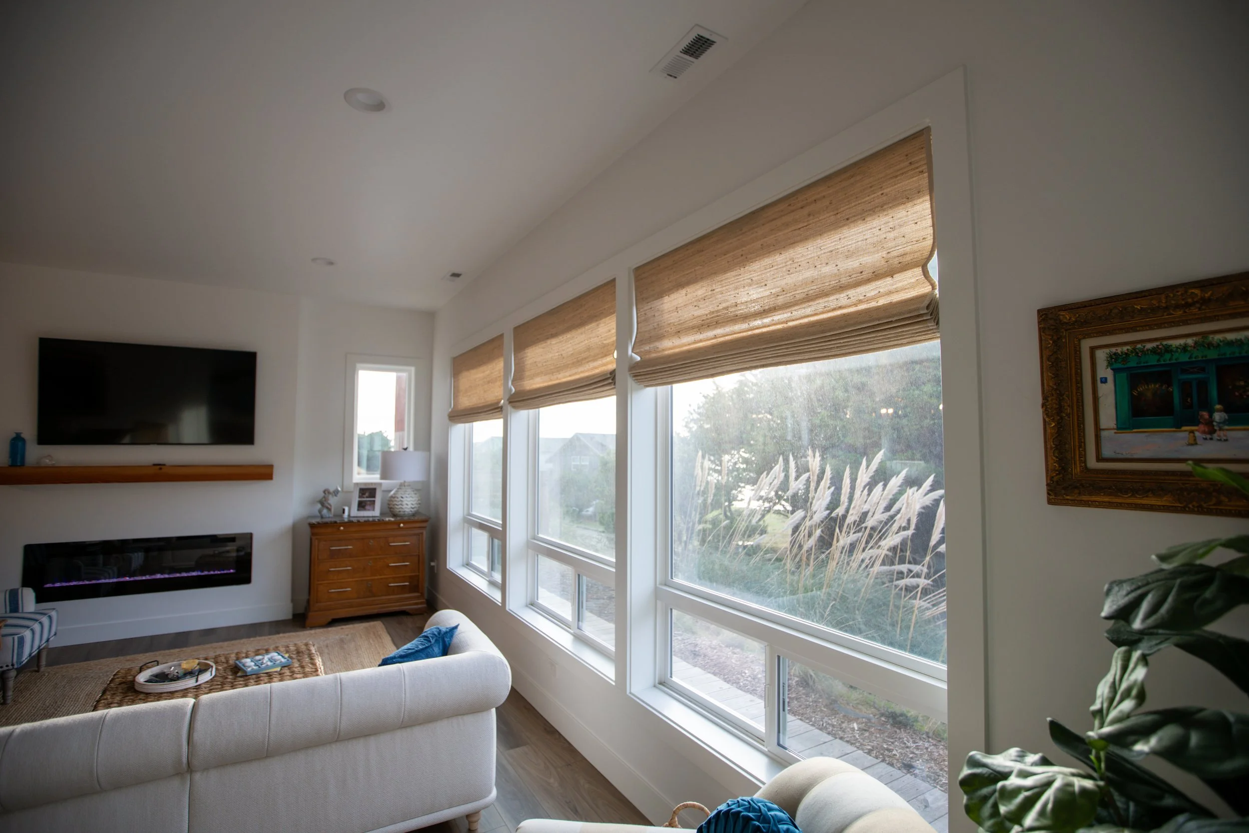 Living room with large windows covered by beige Roman shades, a white sofa, a wooden chest of drawers with a lamp and picture, an electric fireplace, a wall-mounted TV, and a framed painting.