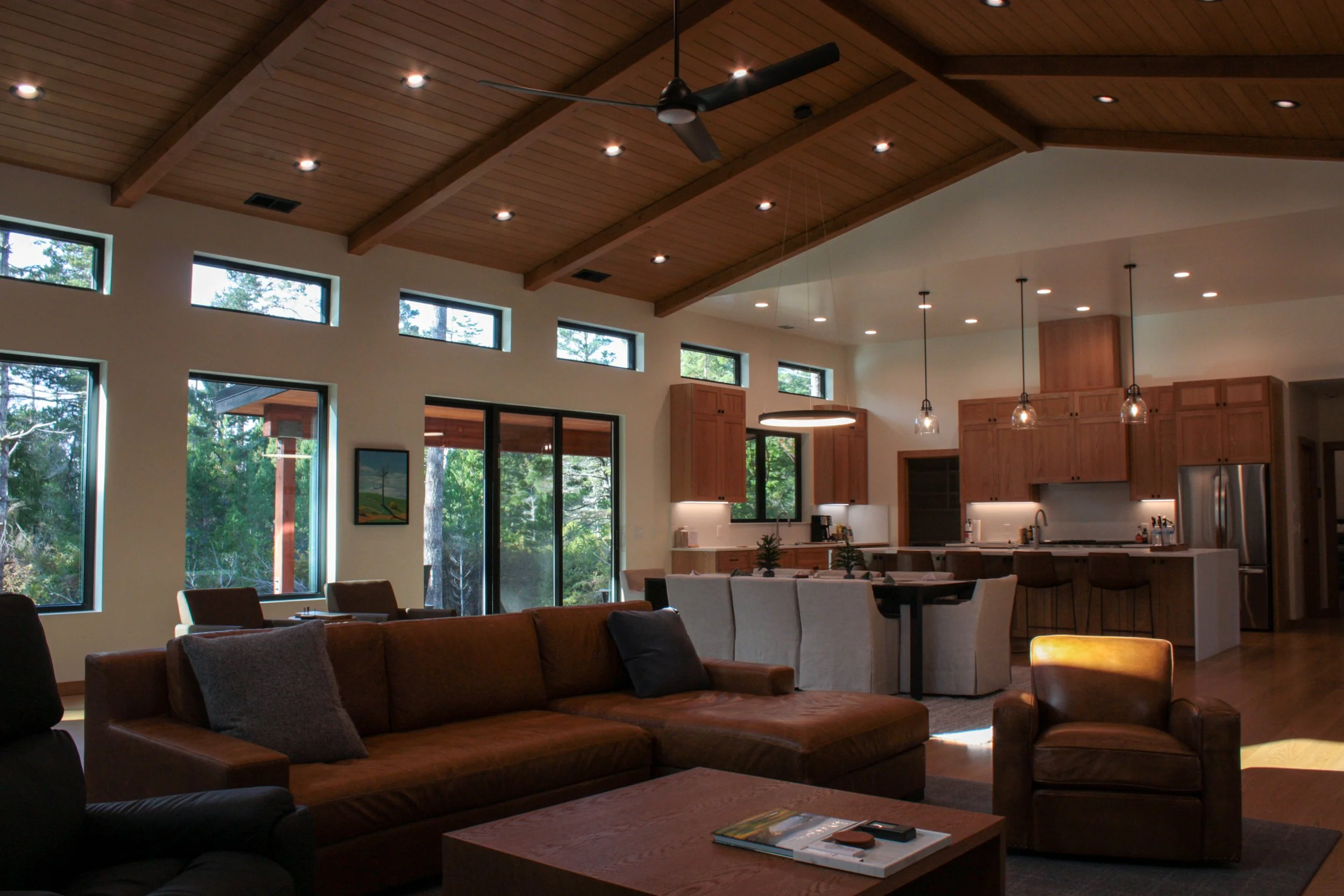 Interior of a modern open-concept living room and kitchen with high wooden vaulted ceiling, multiple windows, a leather sofa, an armchair, and a kitchen with wooden cabinets, island, and pendant lighting.