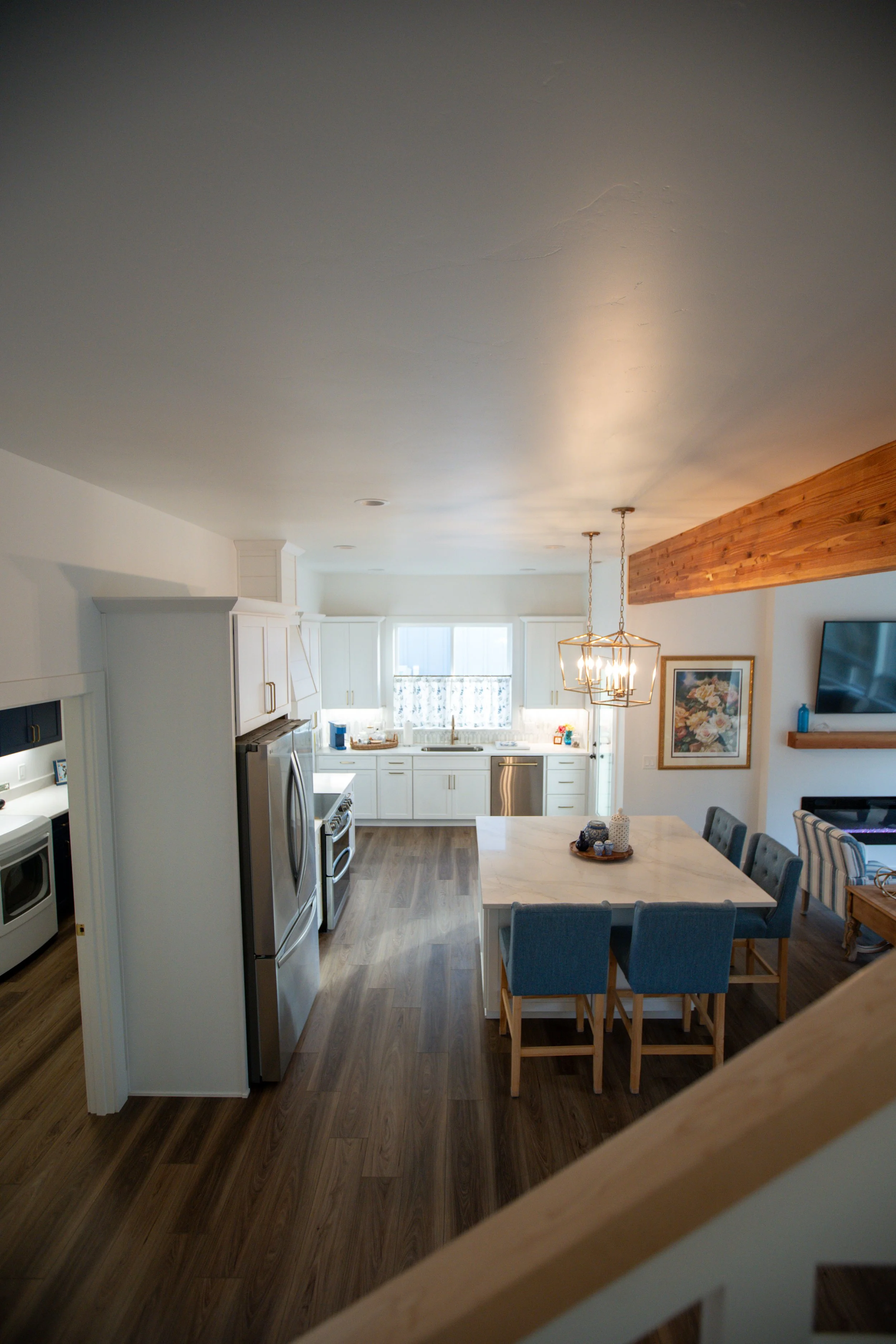 View of a bright, modern kitchen with white cabinets, wooden flooring, and a central island with blue chairs. Natural light fills the space from large windows, and there's a hanging chandelier above the island.
