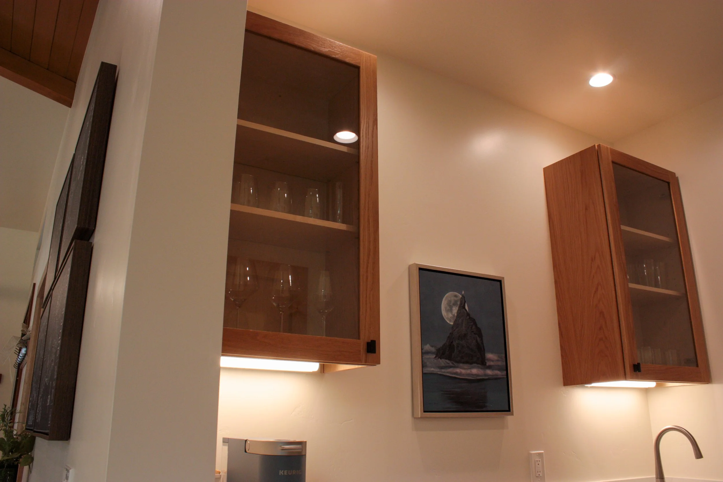 A kitchen wall with two wooden cabinets, a framed moonlit ocean scene painting, and a lemon-shaped light fixture.