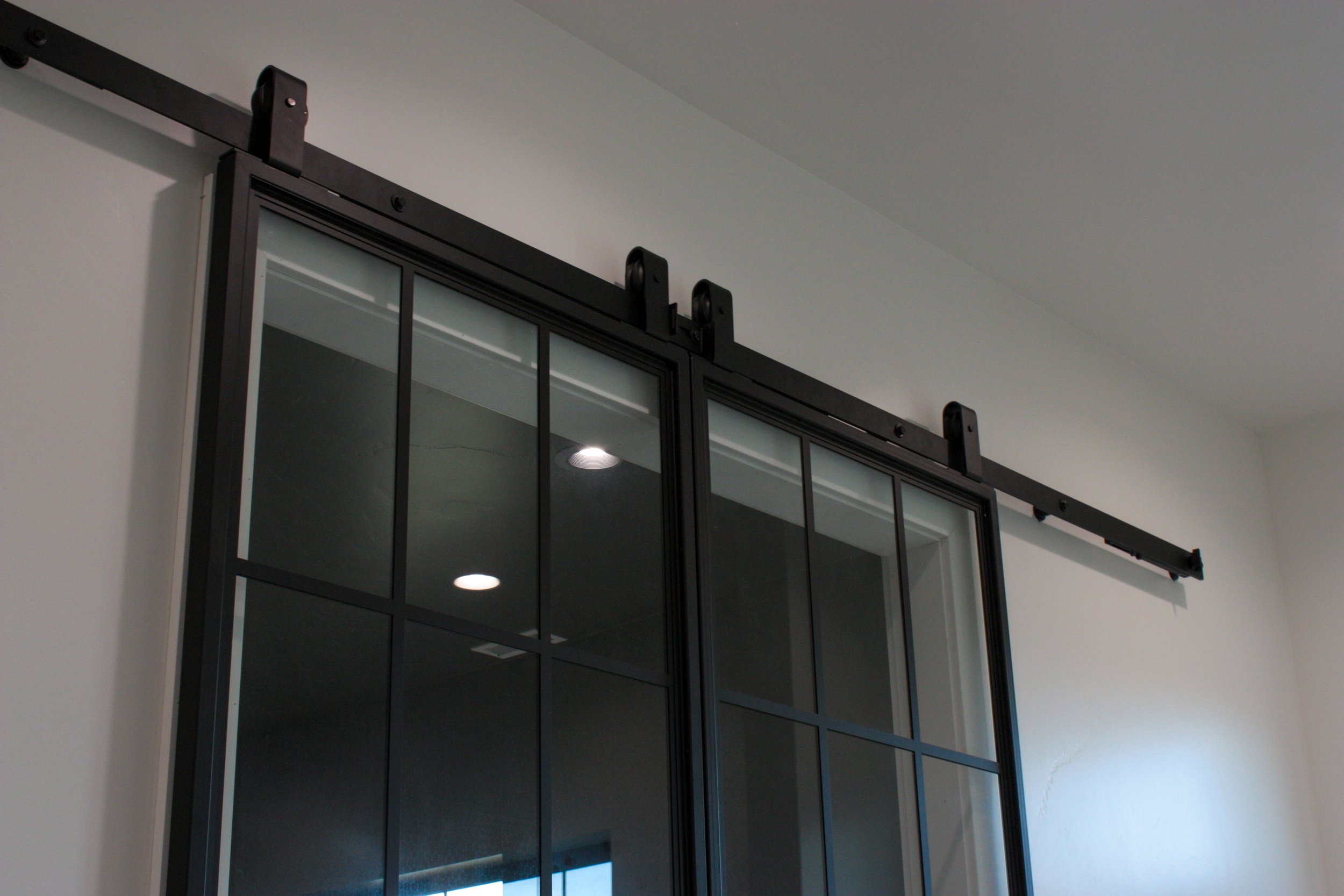 Black sliding barn door with glass panels installed on a white wall, roller hardware mounted on a ceiling track.