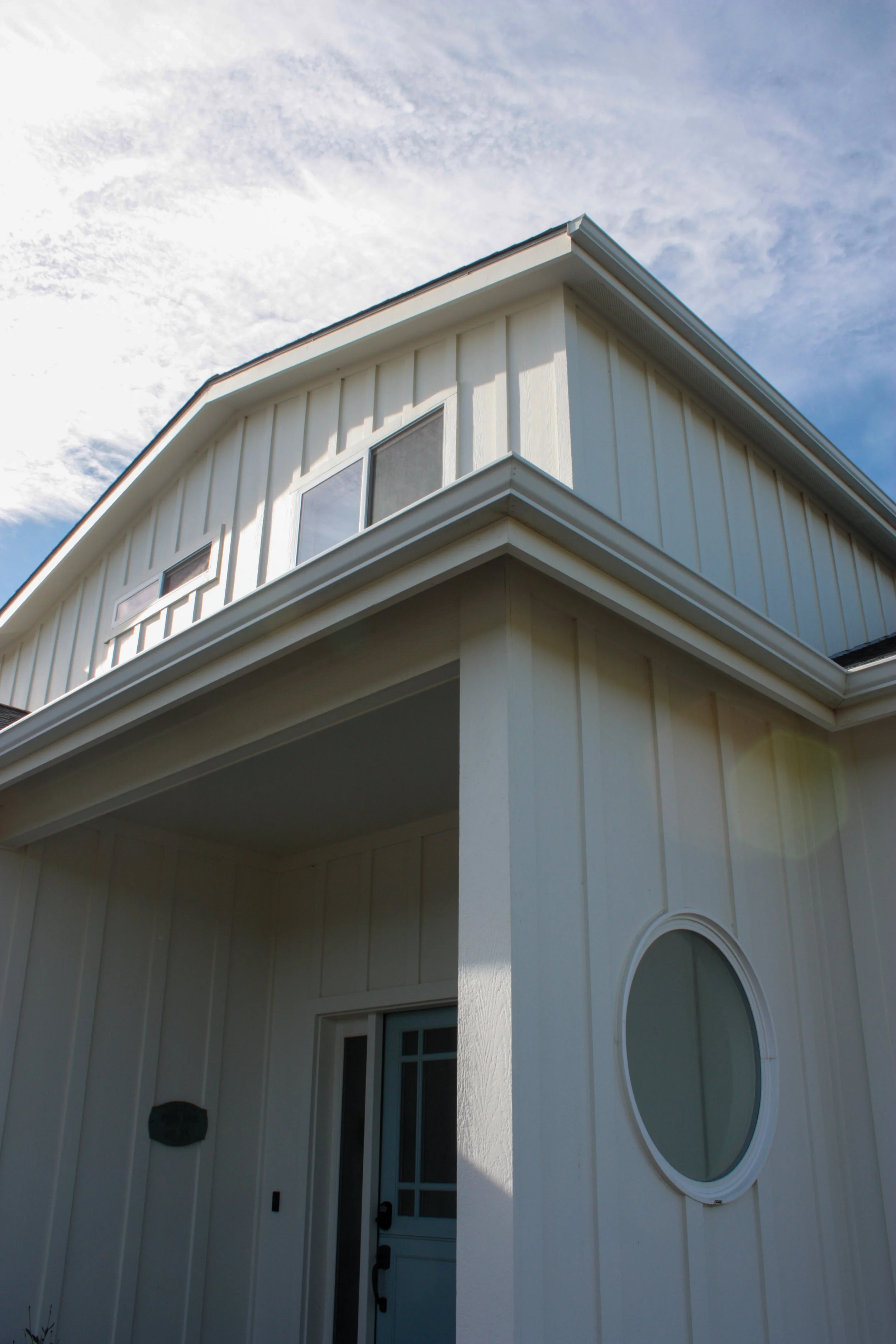 Close-up of a white two-story house with vertical paneling, a corner column, and a round window, under a partly cloudy sky.