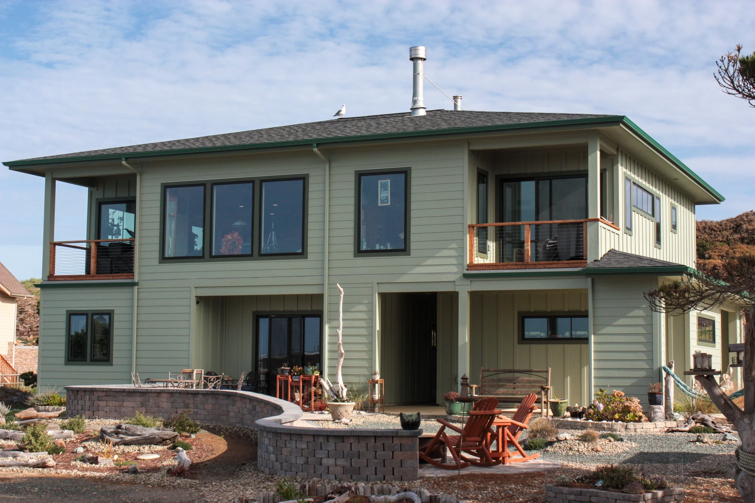 Two-story house with green painted exterior and large windows, small balcony on the upper left, backyard with wooden chairs, gravel and garden plants, clear sky with clouds