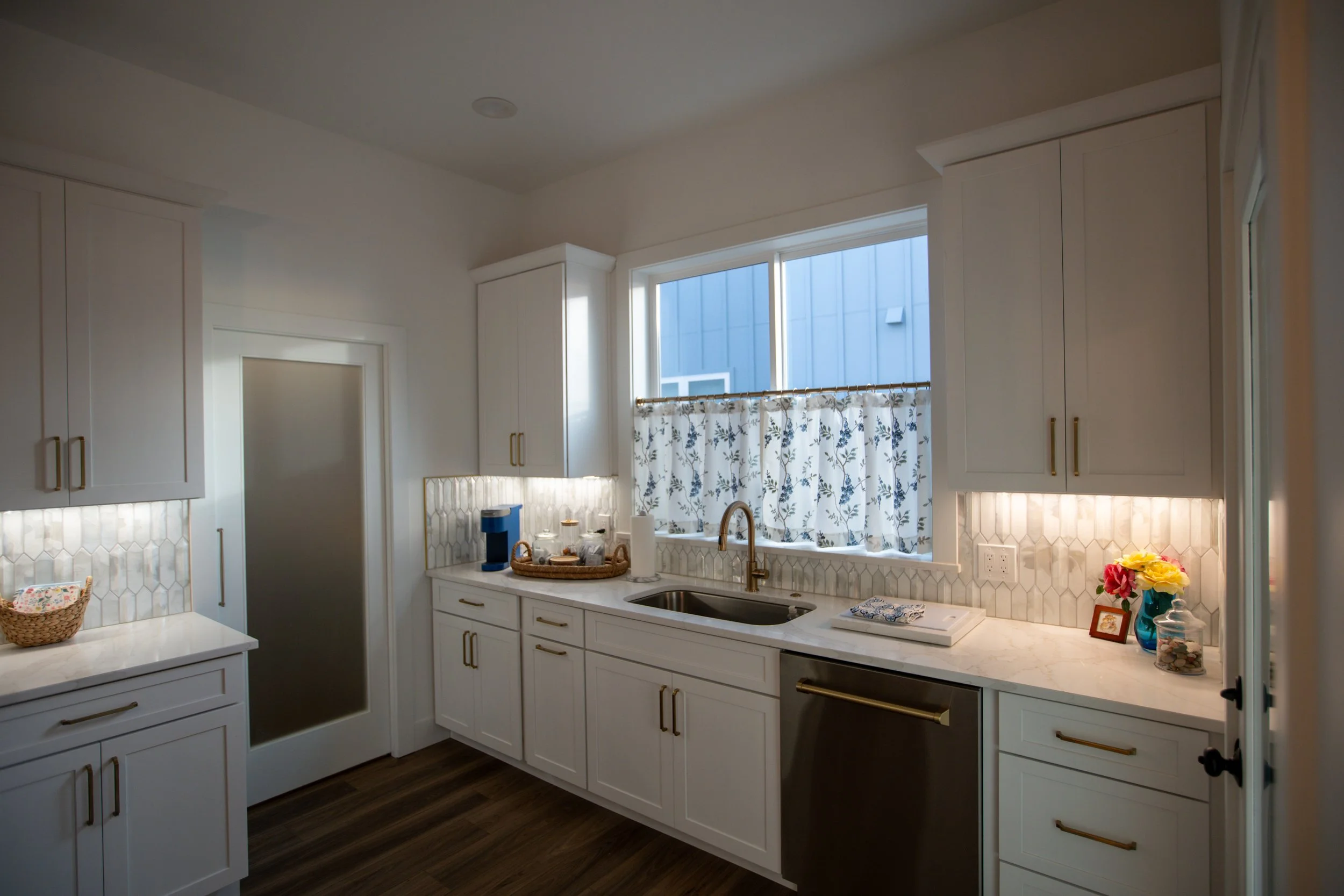 Bright kitchen with white cabinets, marble countertops, a window with floral curtains, and a sink with a gold faucet. Decor includes a vase of flowers, a framed photo, and small jars, with a wood floor.